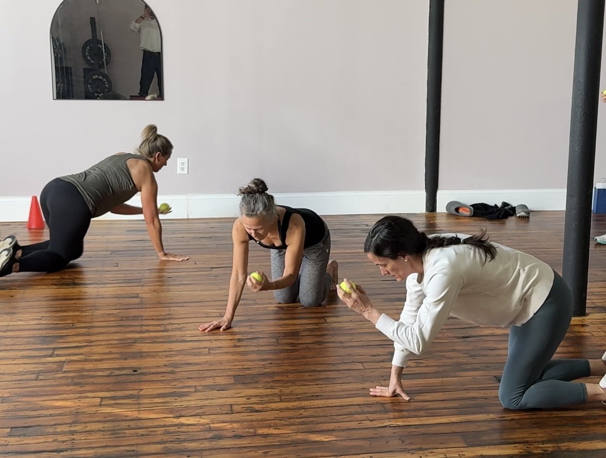 Three women in a fitness class on a wooden floor practicing yoga or stretching exercises, holding yellow balls, with one woman in a white hoodie and gray leggings, another in a black tank top and gray pants, and a third in a dark tank top and black pants.