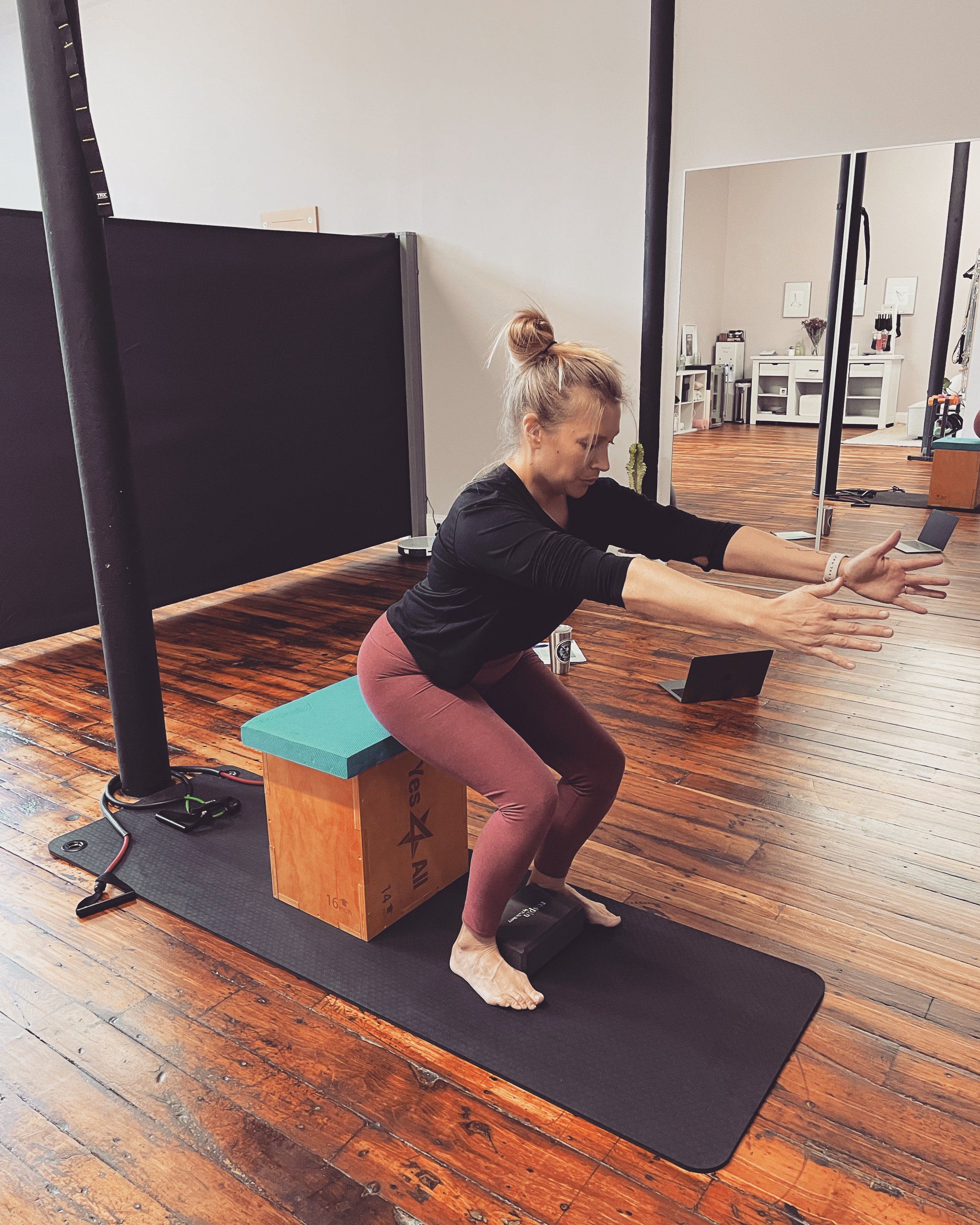 Woman performing a squat exercise on a black mat, sitting on a wooden plyometric box, in a gym with wooden floors and mirrors.