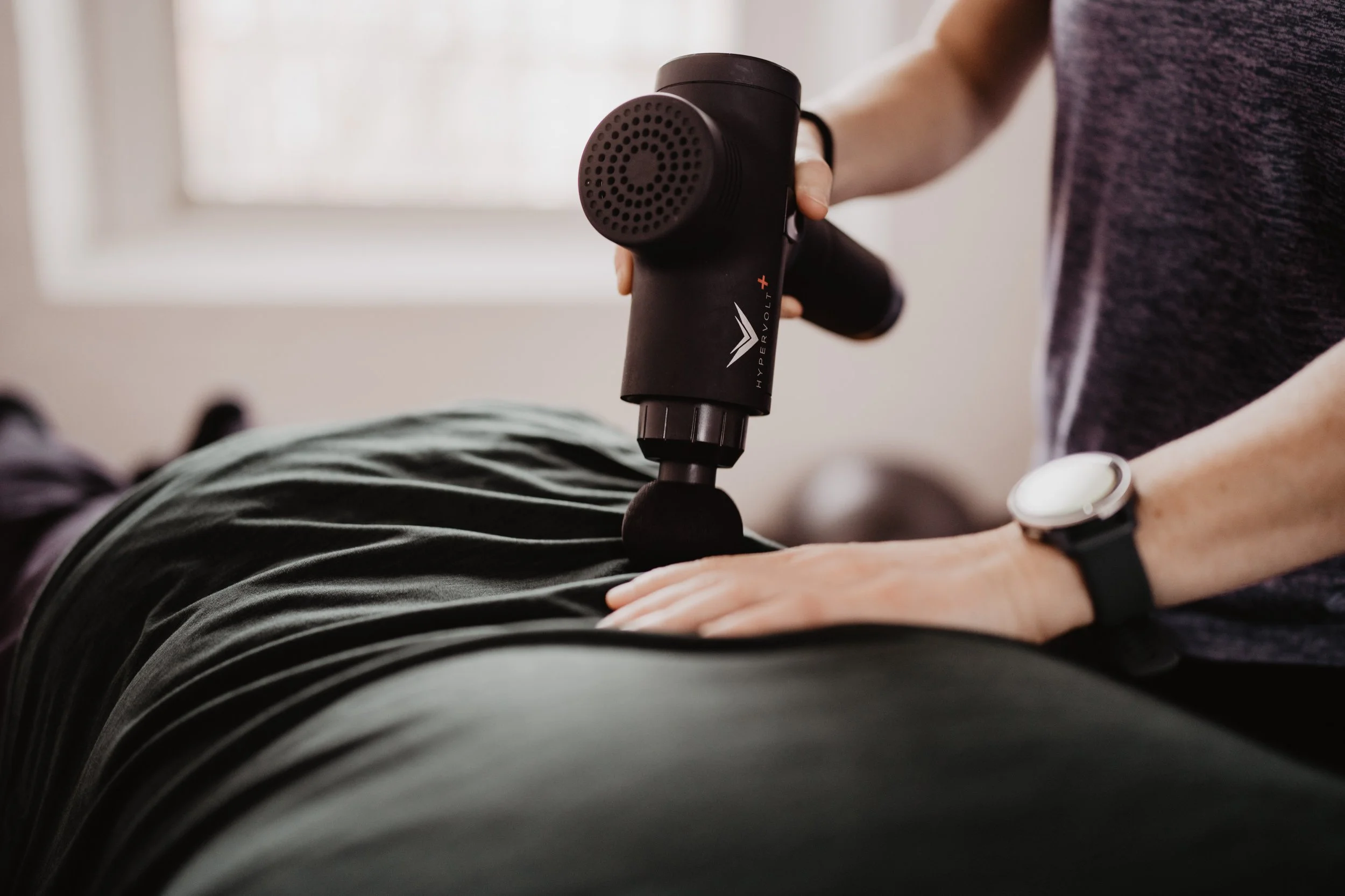 Person using a massage gun on a patient's lower back in a therapy setting.
