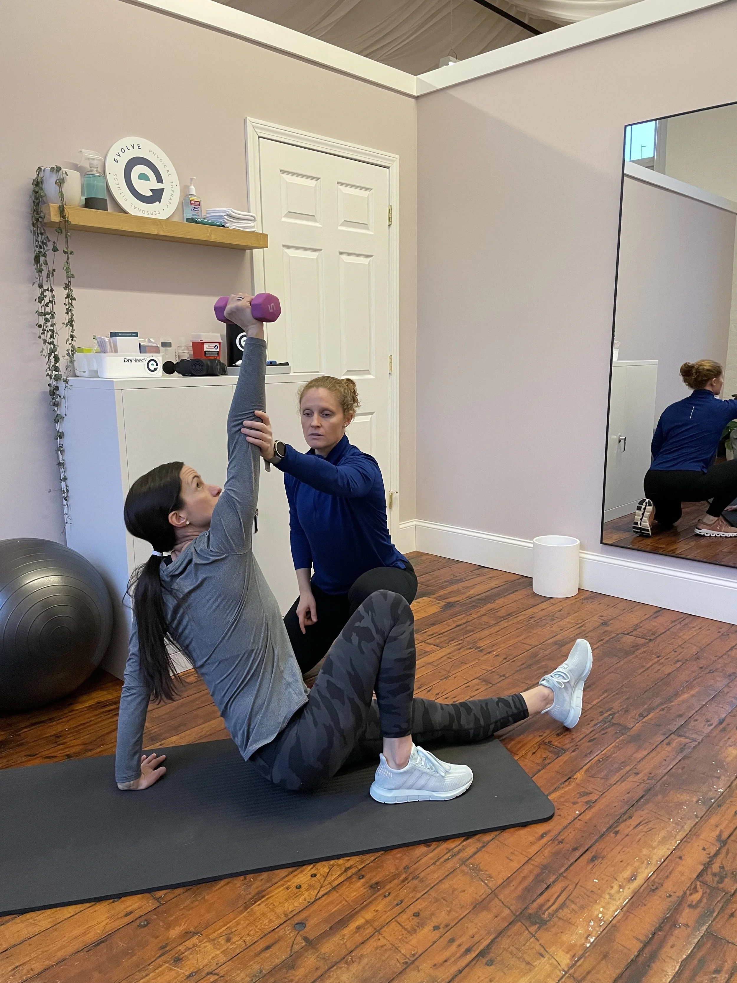 A woman sitting on a yoga mat doing a workout with a trainer helping her lift a pink dumbbell overhead in a fitness studio.