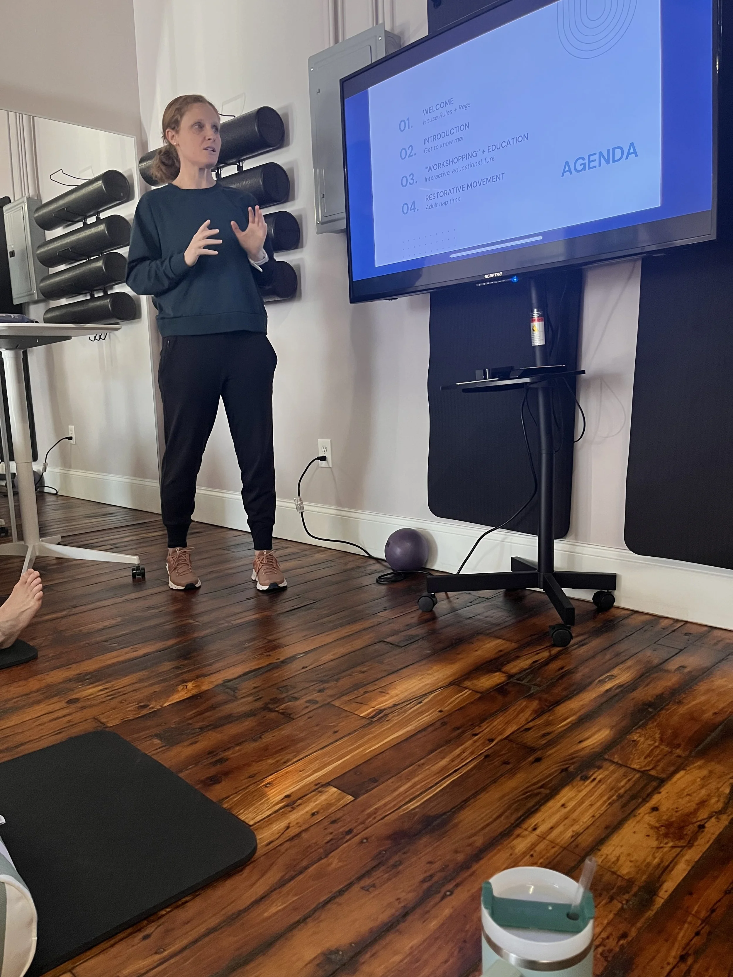 A woman giving a presentation in a fitness or wellness setting with a large screen displaying an agenda, including welcome, introduction, 'workshopping' education, and restorative movement.