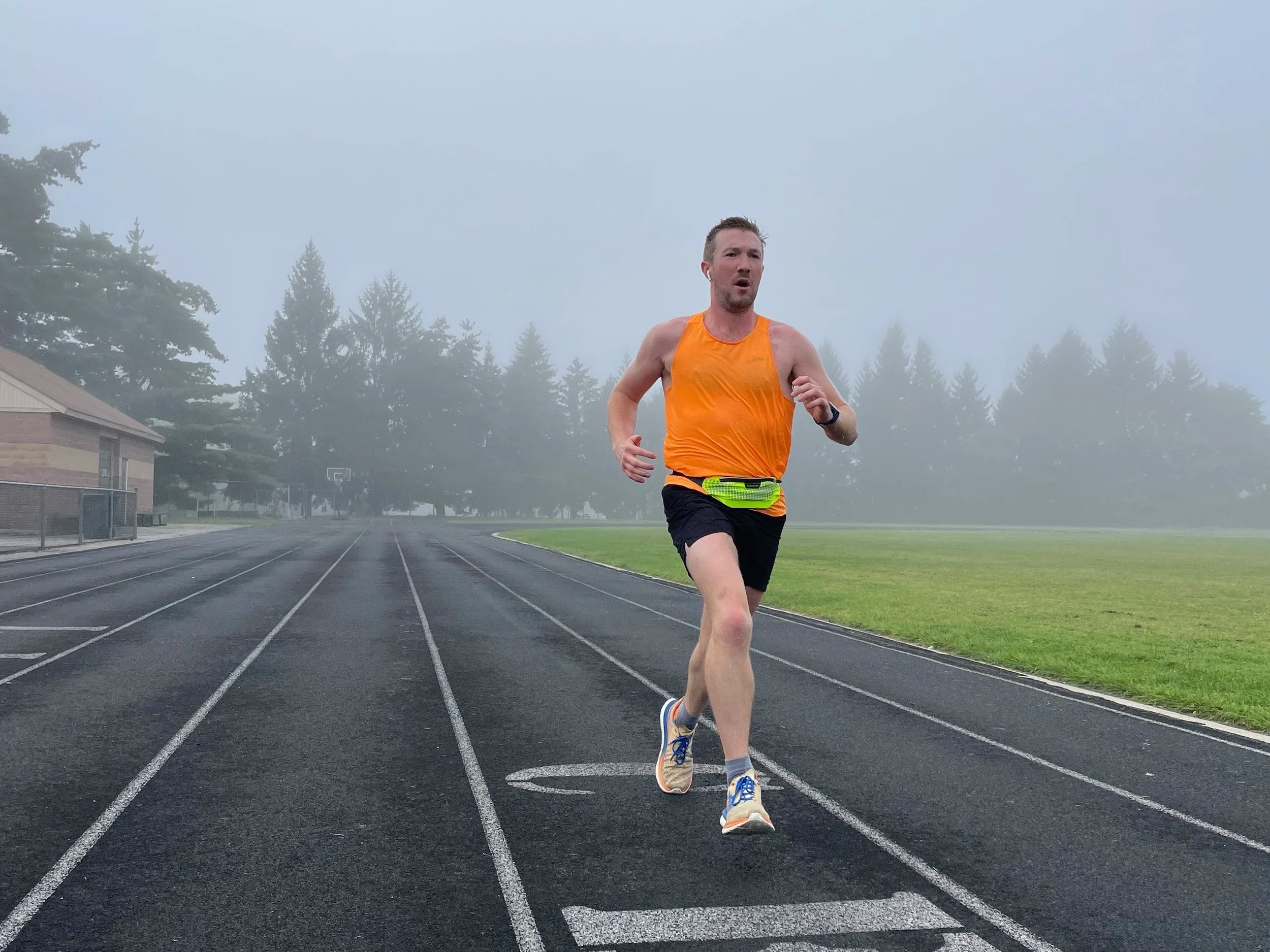 A man running on a track field during foggy weather, wearing an orange tank top, black shorts, and running shoes, with trees and a building in the background.