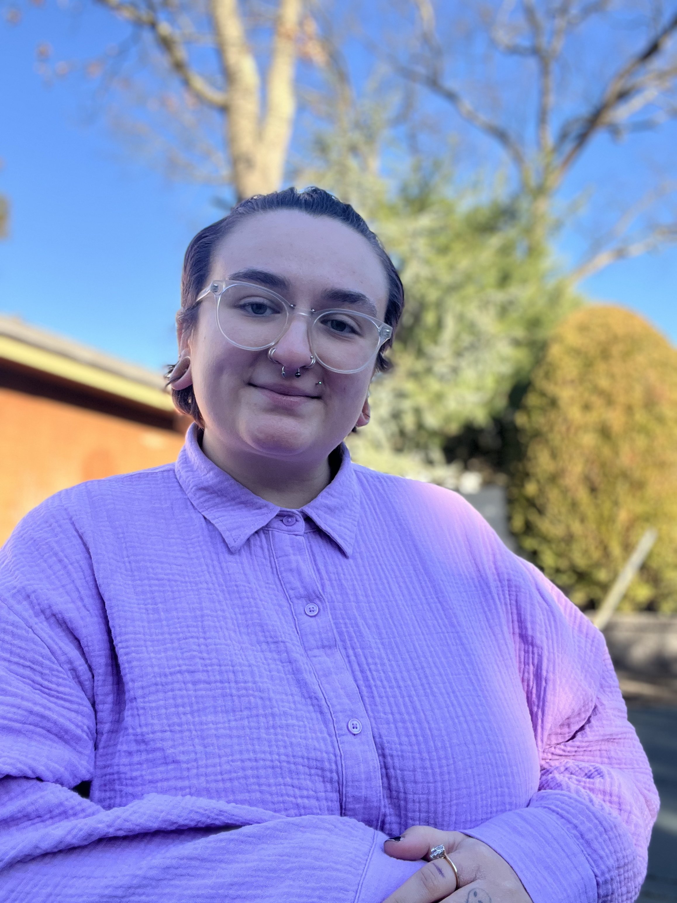 Portrait of a person with short dark hair, wearing glasses, a purple button-up shirt, and multiple nose piercings, standing outdoors with trees and a building in the background.