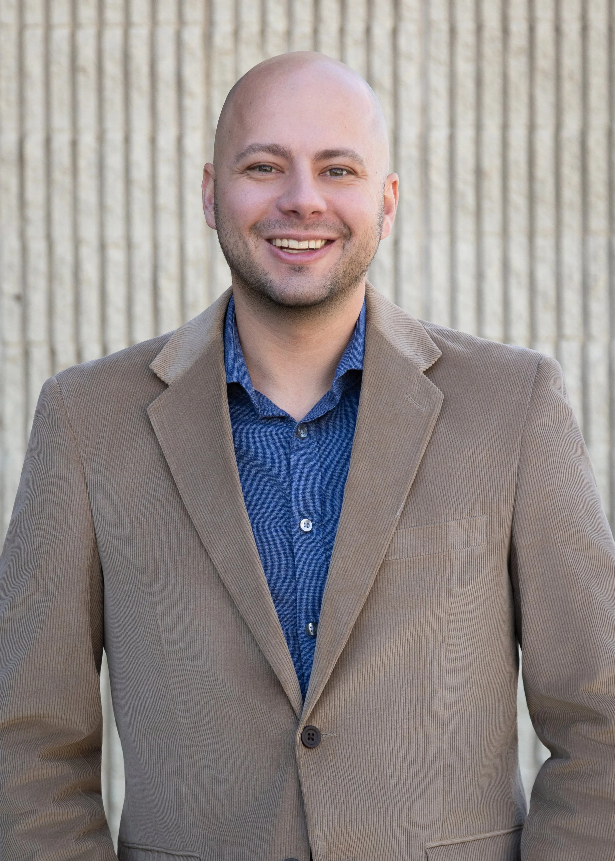A man smiling, wearing a beige blazer over a blue button-up shirt, standing in front of a textured beige wall.