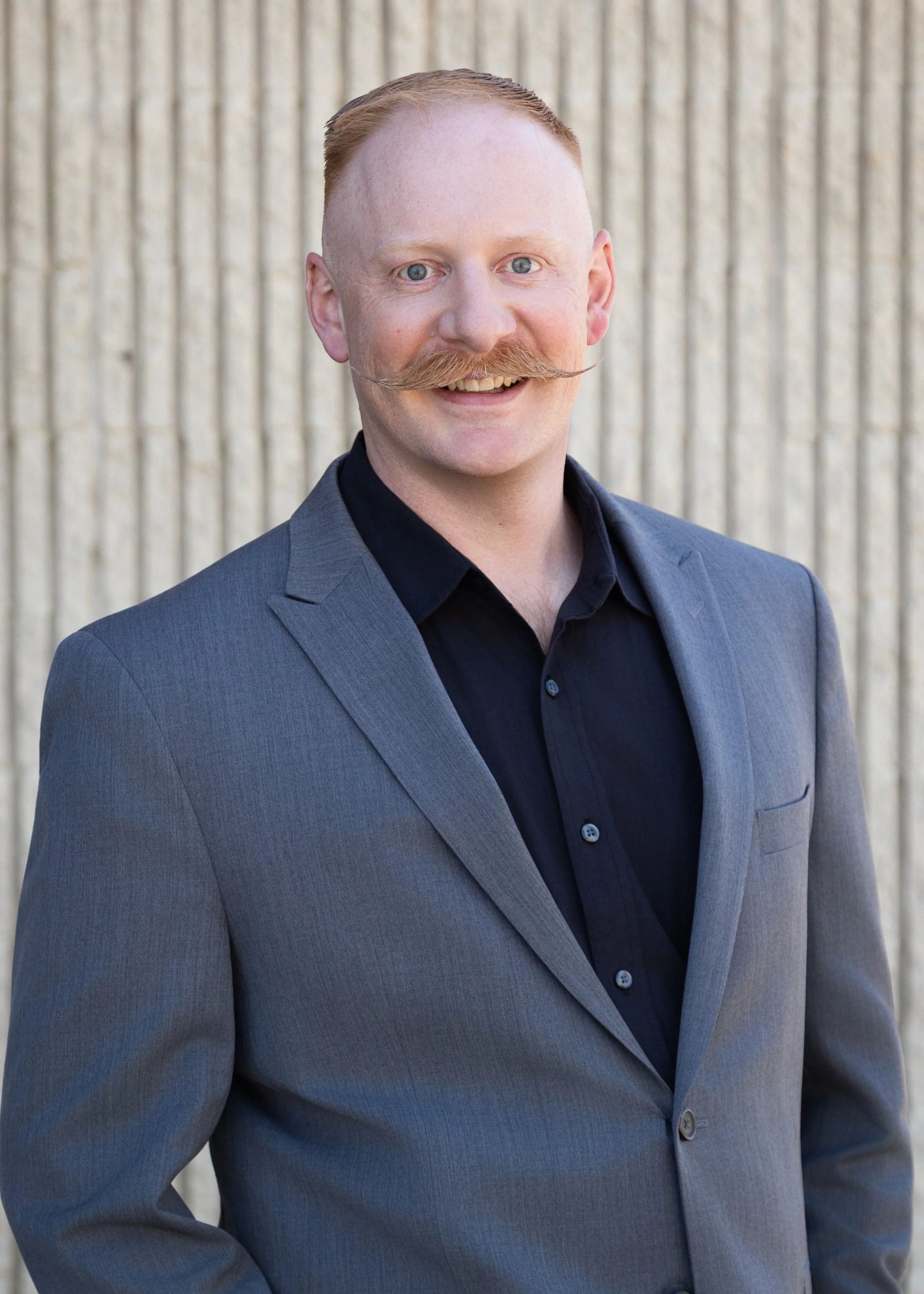 A man with red hair, a large handlebar mustache, and a friendly smile, dressed in a dark shirt and a gray suit, standing in front of a light-colored, vertically ridged wall.
