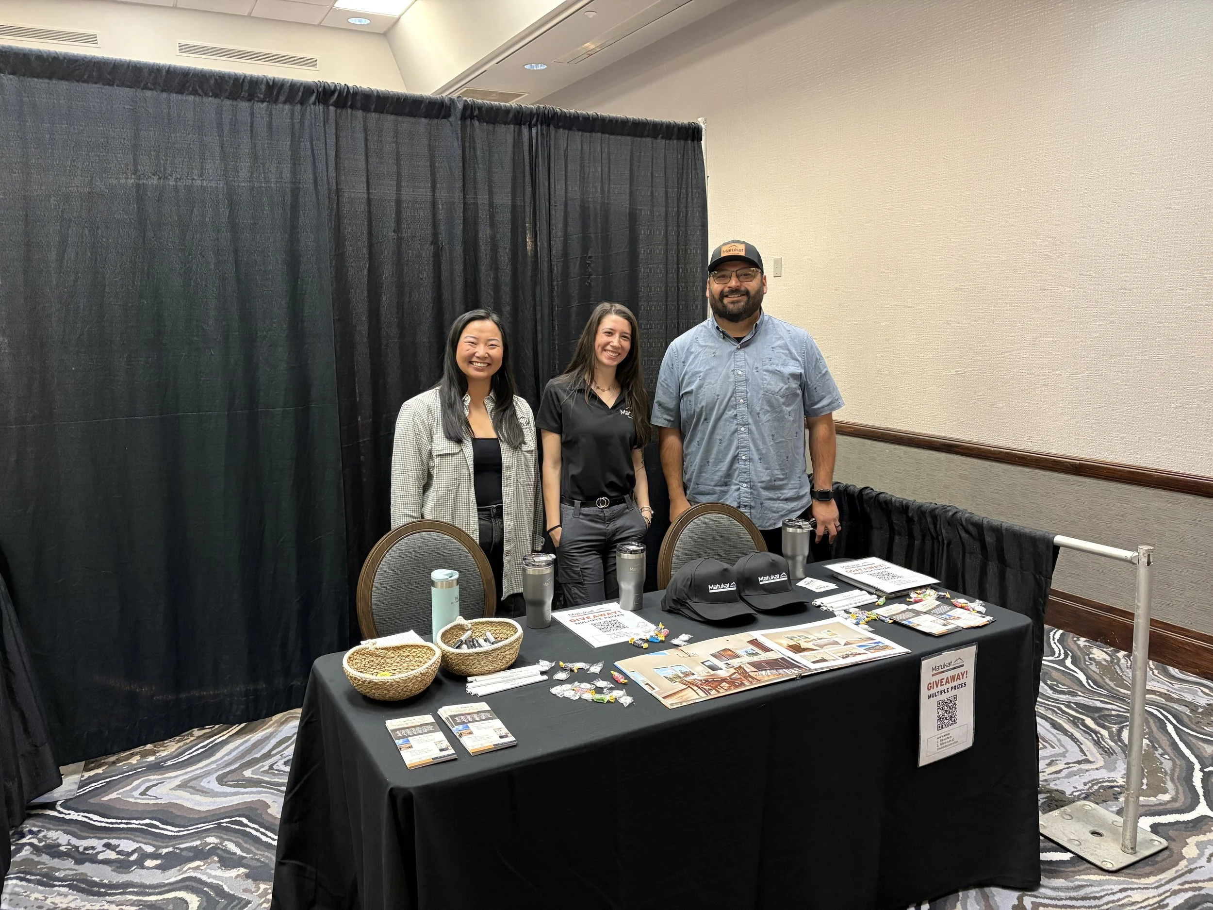Three people standing behind a table with various promotional items at an indoor event. Two women on the left are smiling, and a man on the right is also smiling and wearing glasses and a hat. The table has flyers, small items, and giveaways.