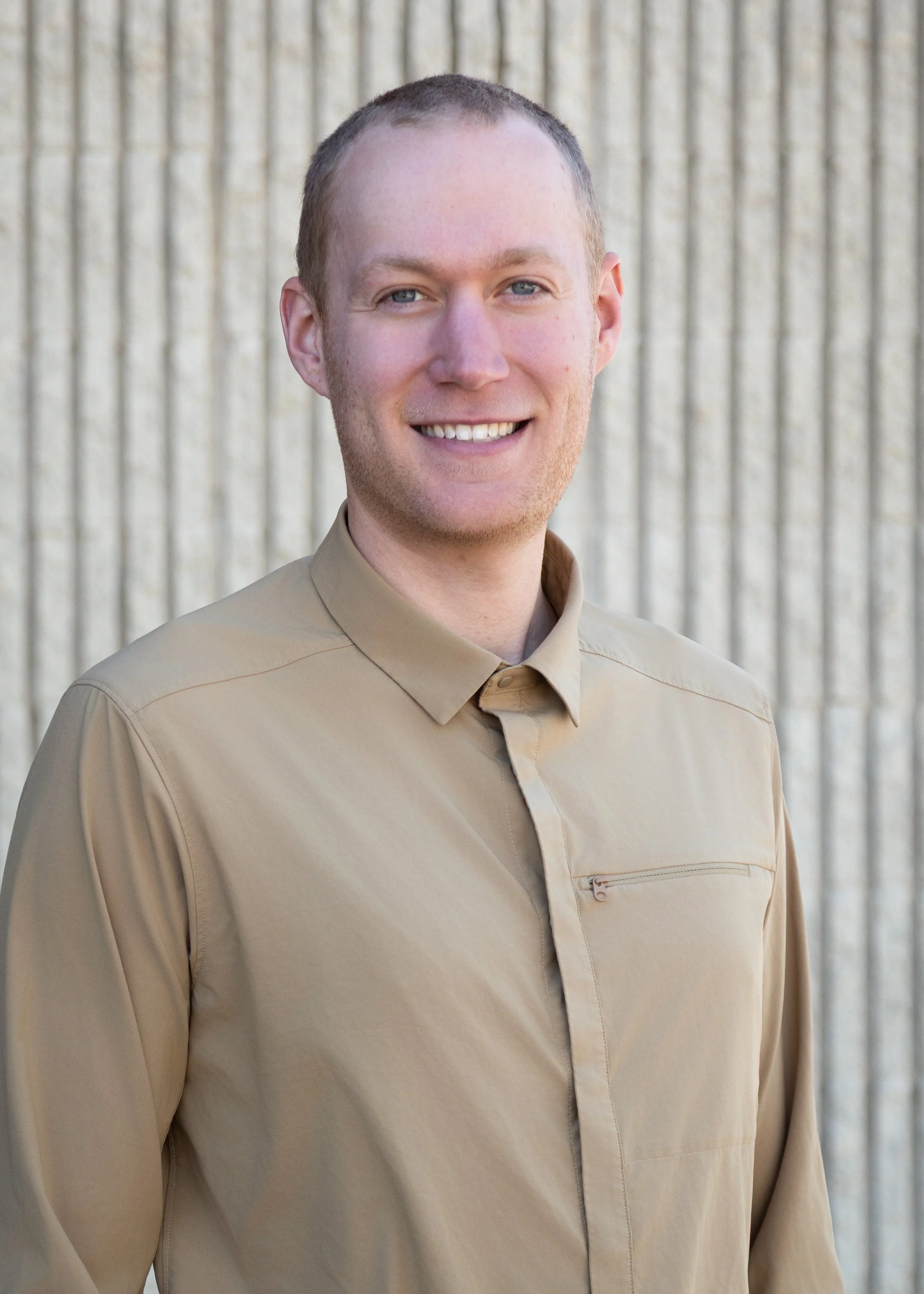 A young man with short hair, blue eyes, and light skin, wearing a tan button-up shirt, smiling, standing outdoors in front of a textured beige wall.