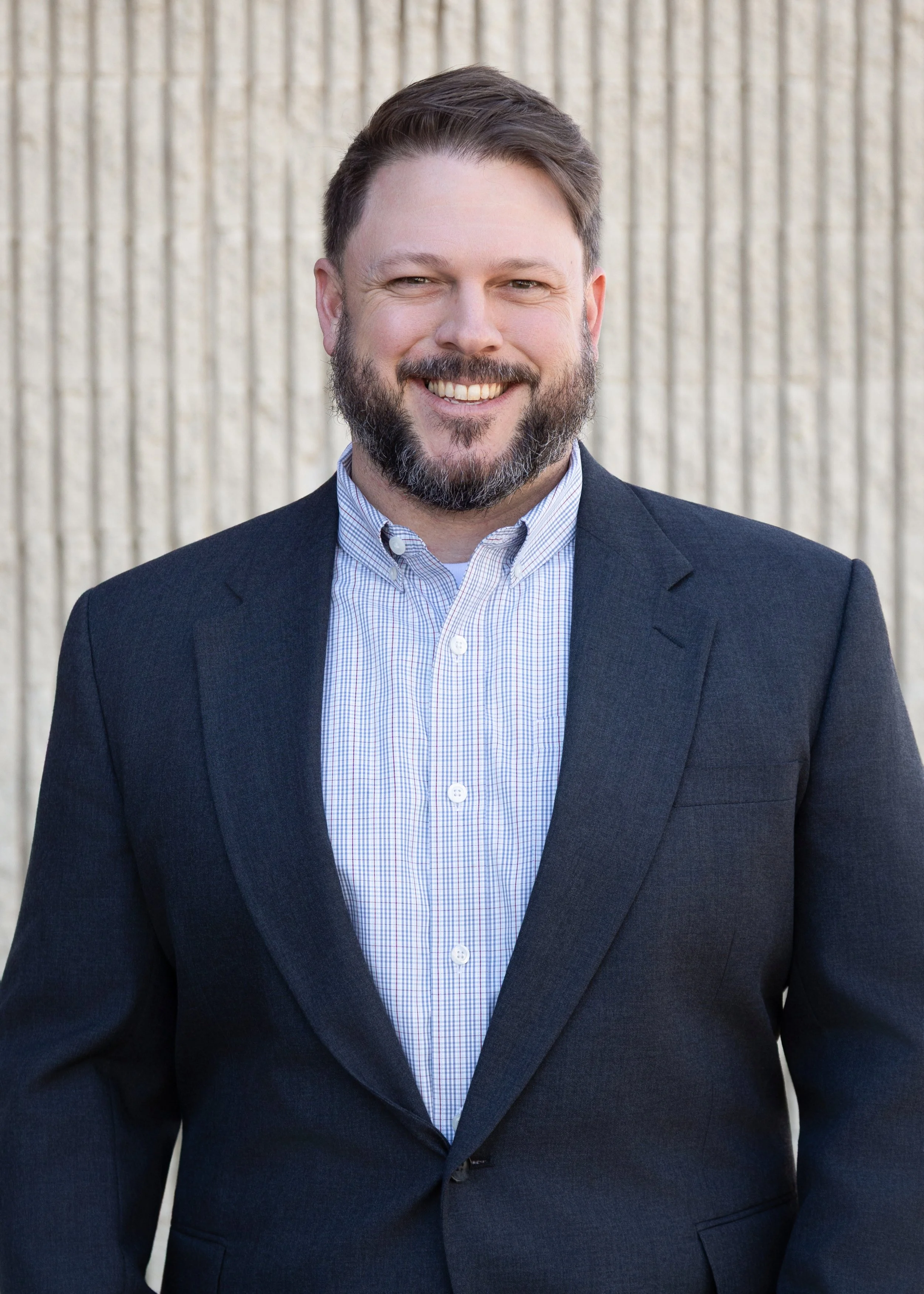A man with a beard smiling, wearing a suit jacket and a collared shirt, standing against a textured wall background.
