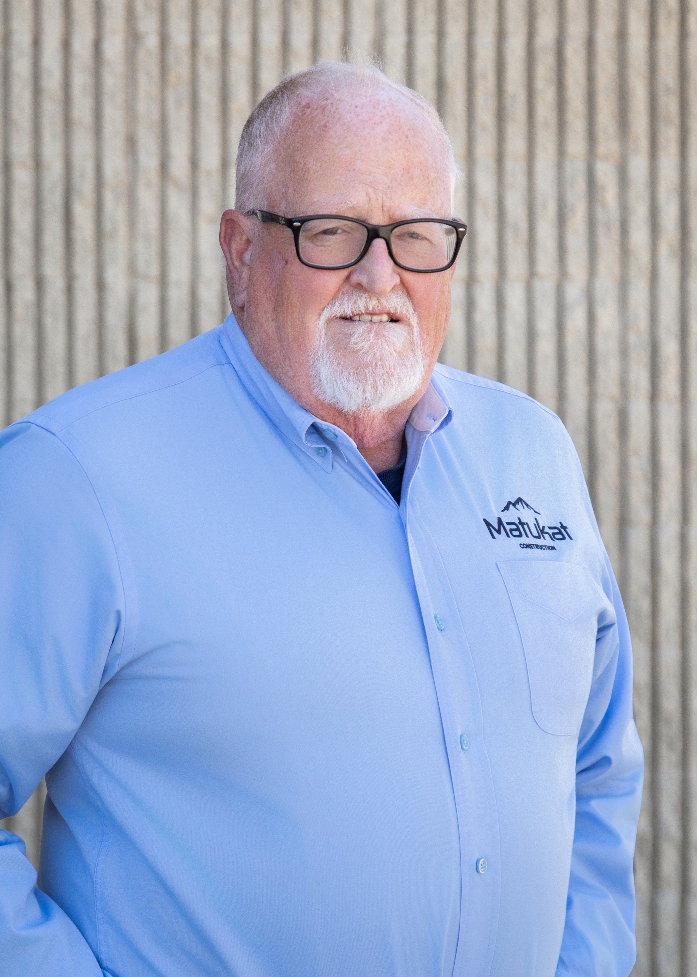 A smiling elderly man with white hair, a beard, and glasses wearing a light blue button-up shirt with a logo on the chest that reads 'Matukat Construction,' standing outdoors in front of a vertical wooden fence.