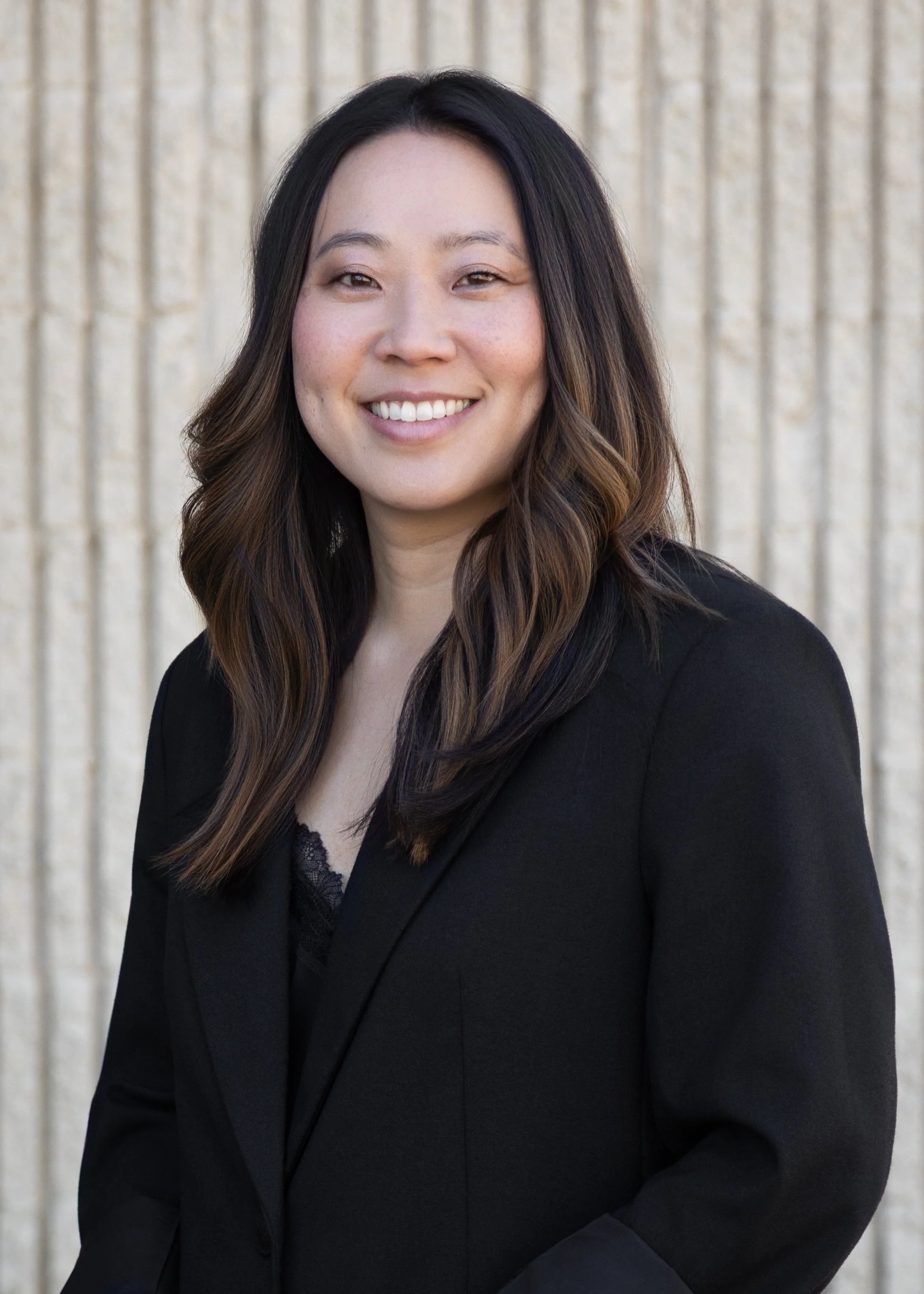 A woman with long dark hair and light makeup, smiling and wearing a black blazer, standing in front of a beige textured wall.