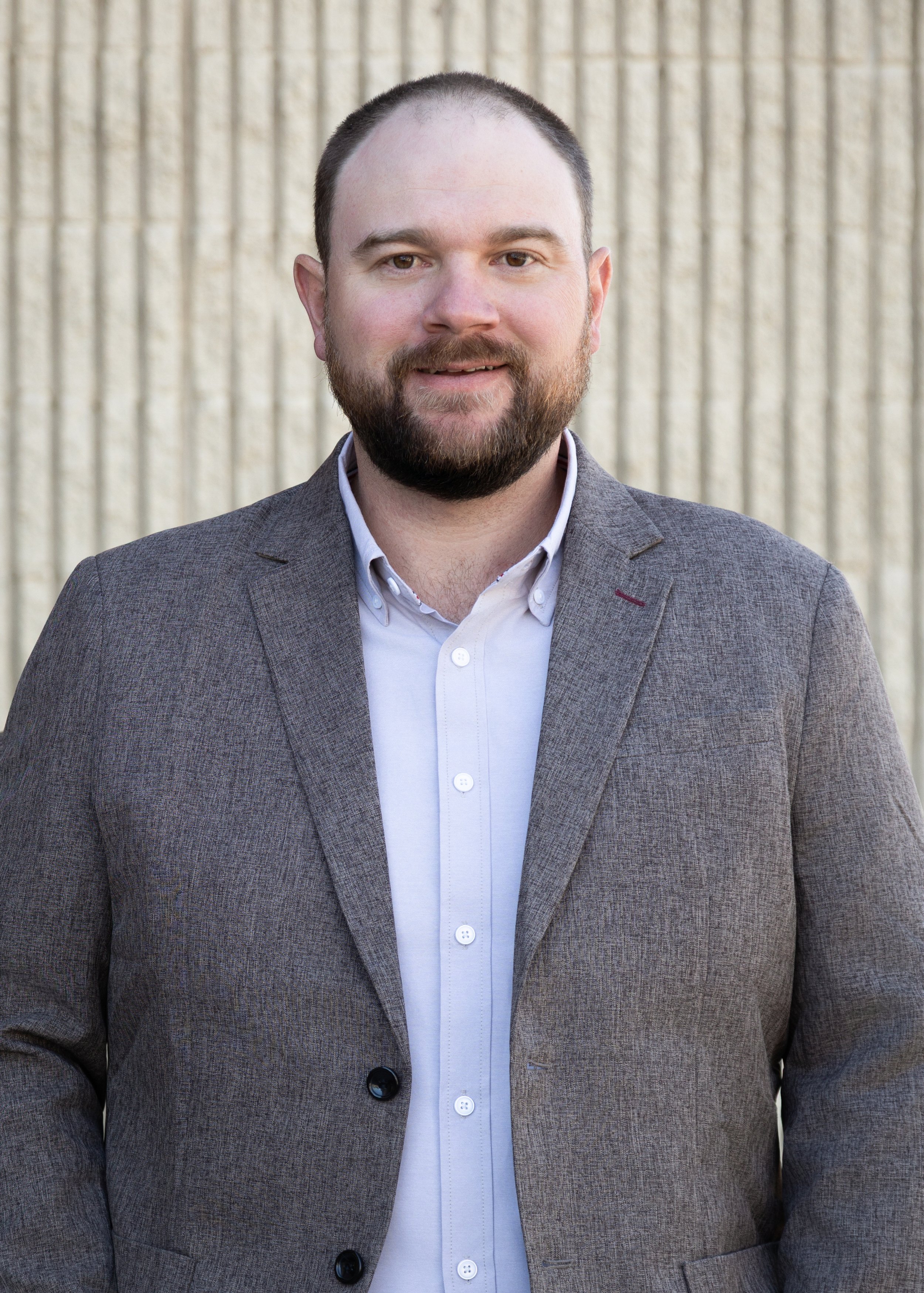 A man wearing a gray blazer and white shirt standing outdoors in front of a beige textured wall.