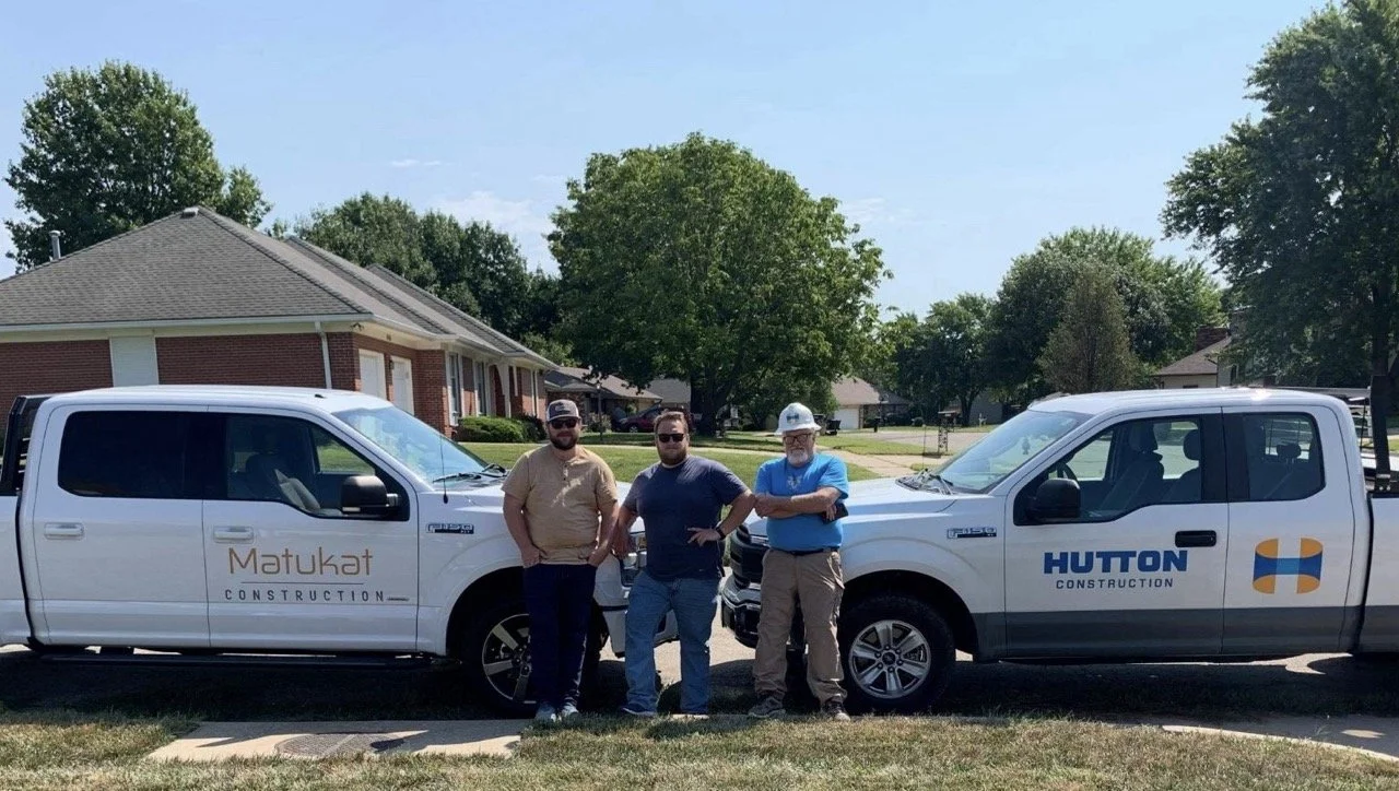 Three construction workers stand in front of two pickup trucks with company logos, on a residential street with houses and trees.