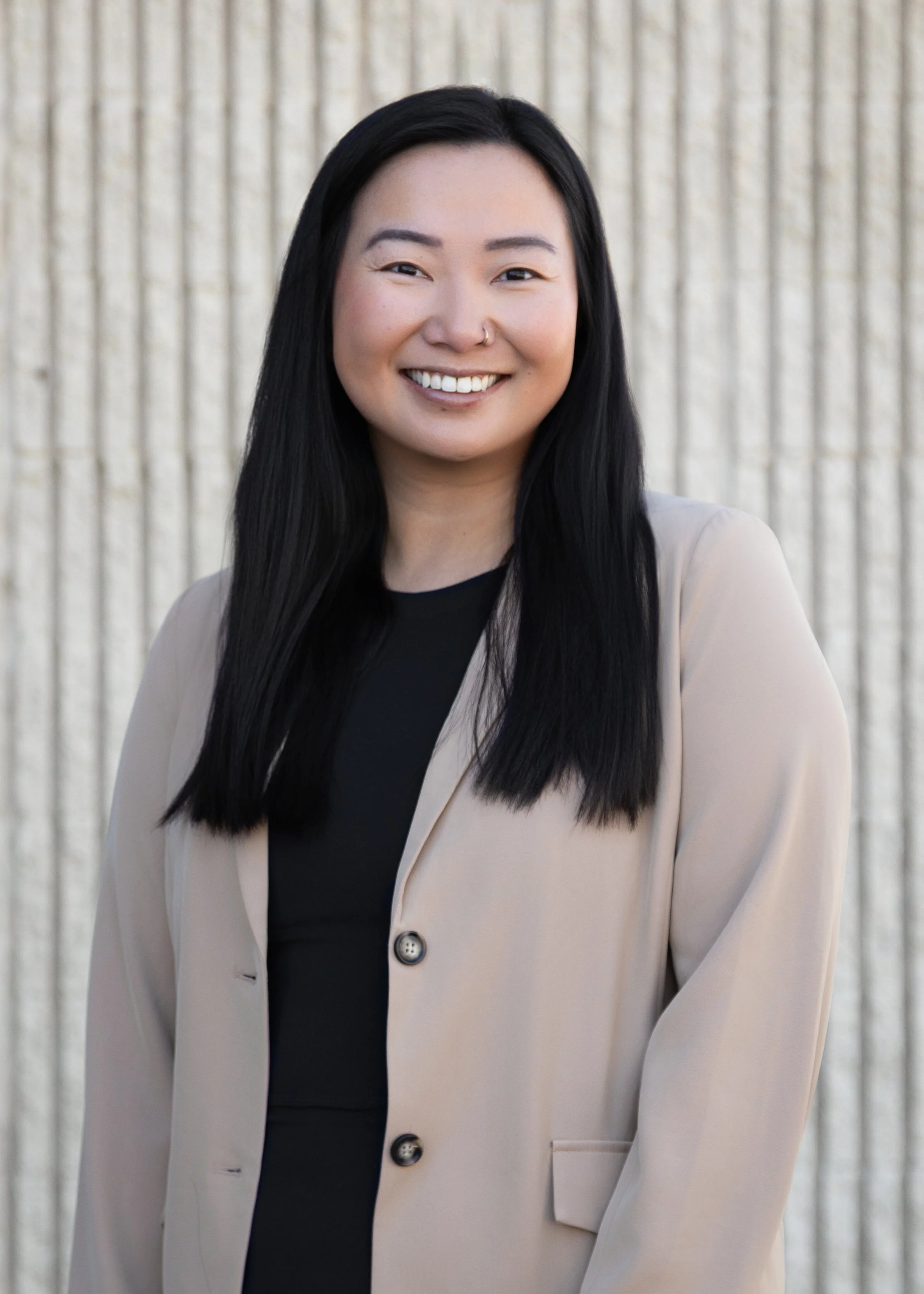 Portrait of a smiling woman with long black hair, wearing a beige blazer over a black top, standing in front of a textured light-colored background.