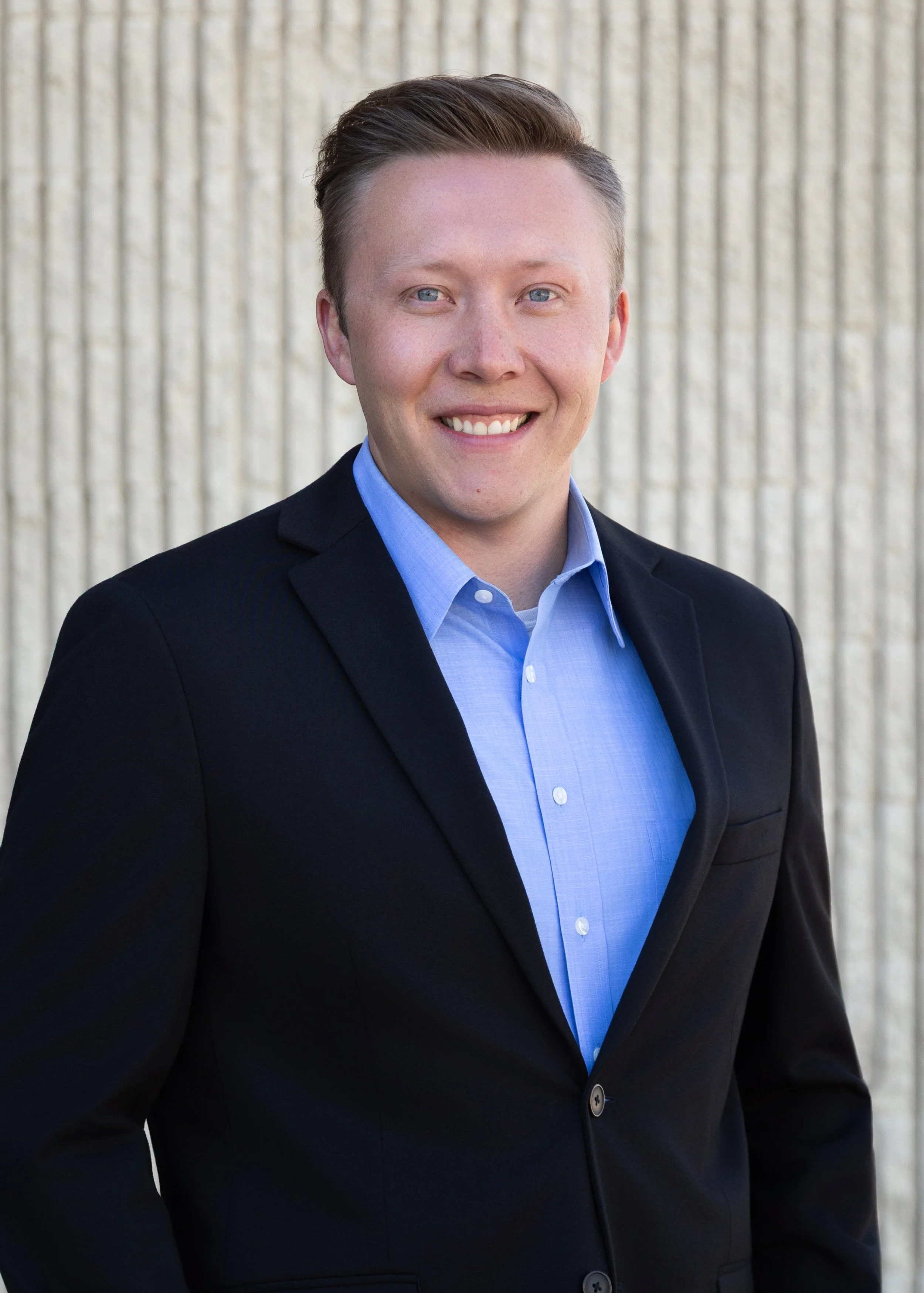 A young man with light skin, light blue eyes, and short, neatly styled brown hair smiling, wearing a light blue dress shirt and a dark blazer, standing outdoors in front of a textured concrete wall.