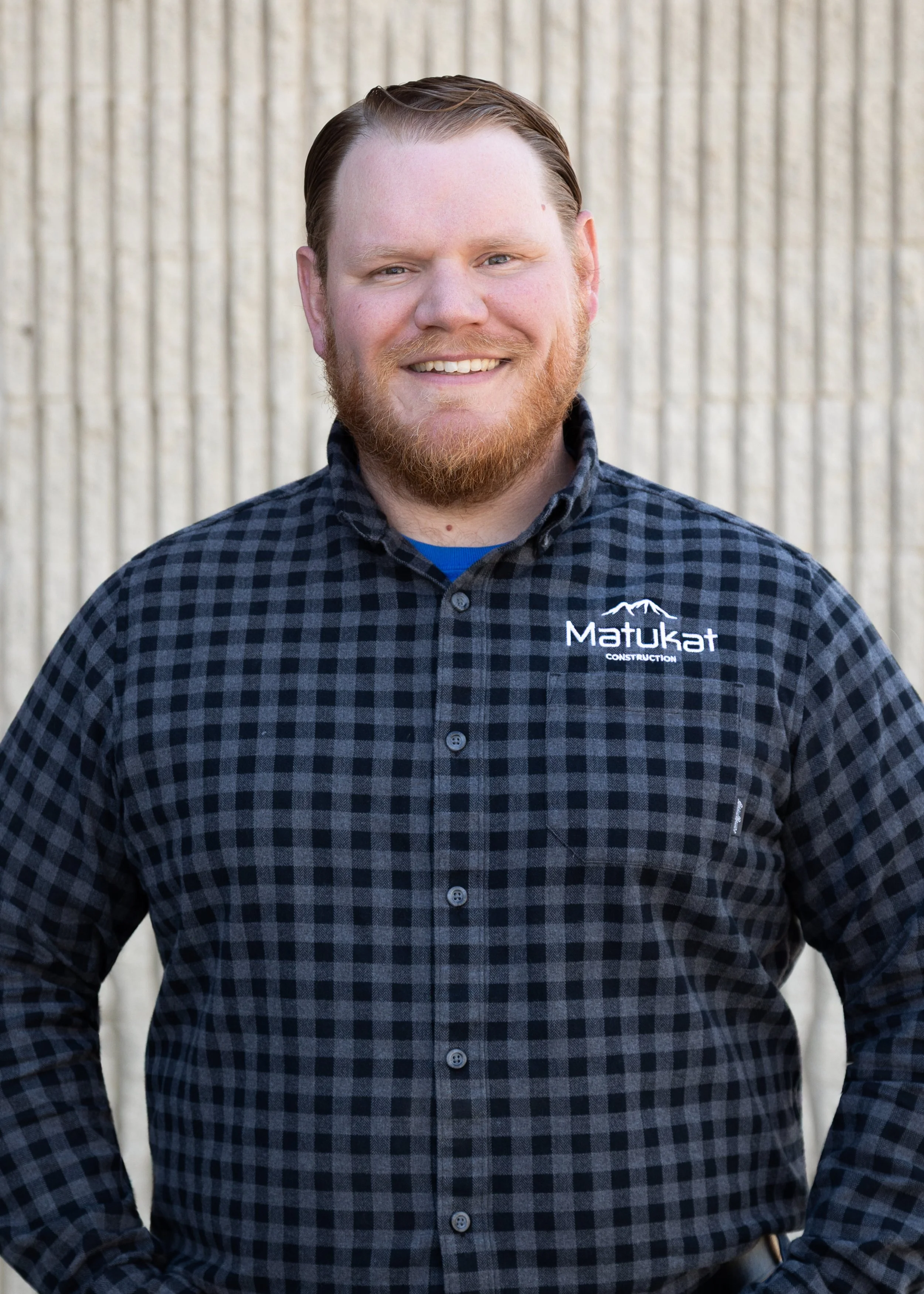 A smiling man wearing a black and gray checkered shirt with a logo that says 'Matukat Construction,' standing outdoors in front of a textured beige wall.