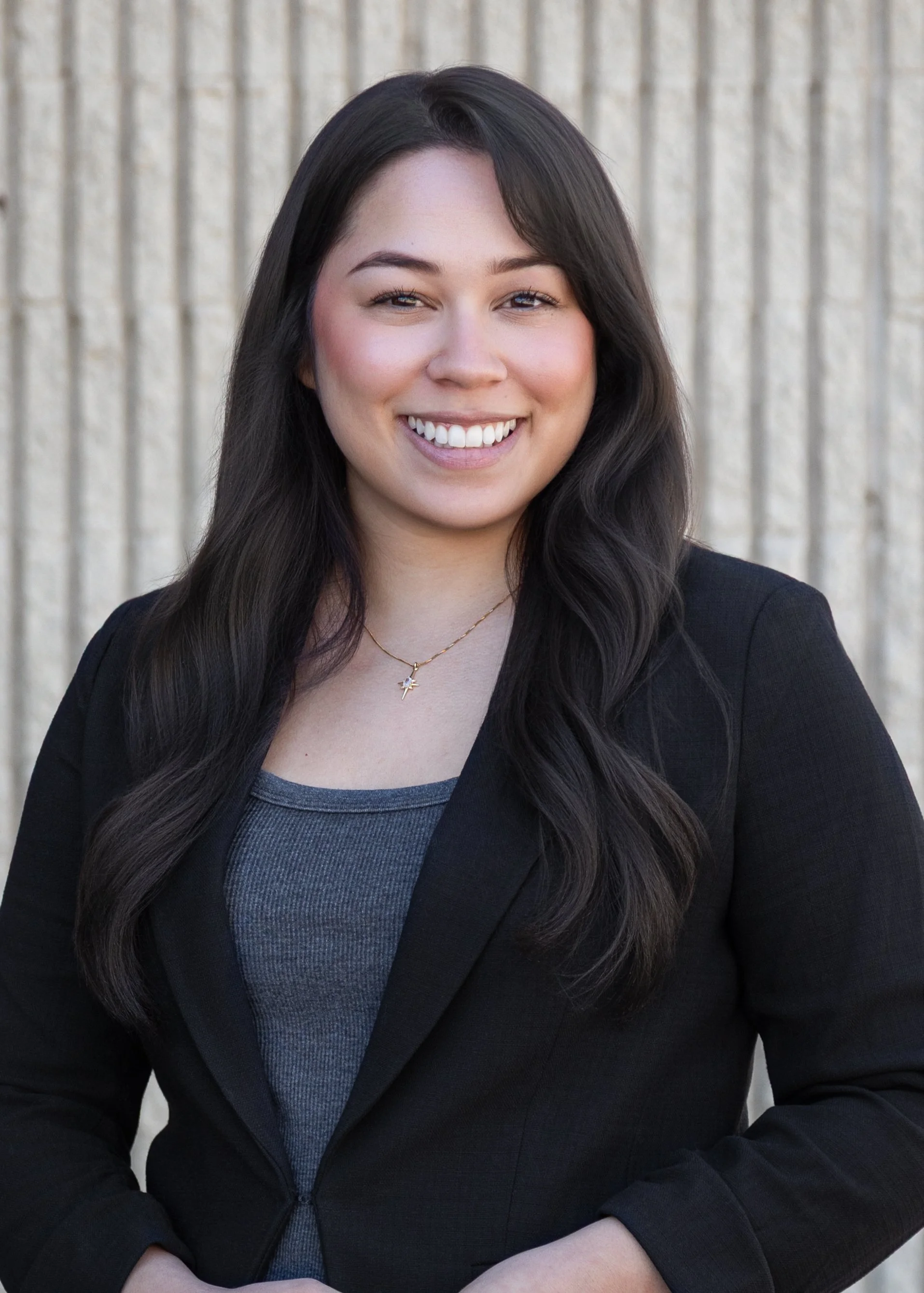 Close-up of a smiling young woman with long dark hair, wearing a black blazer and blue top, standing against a wooden background.