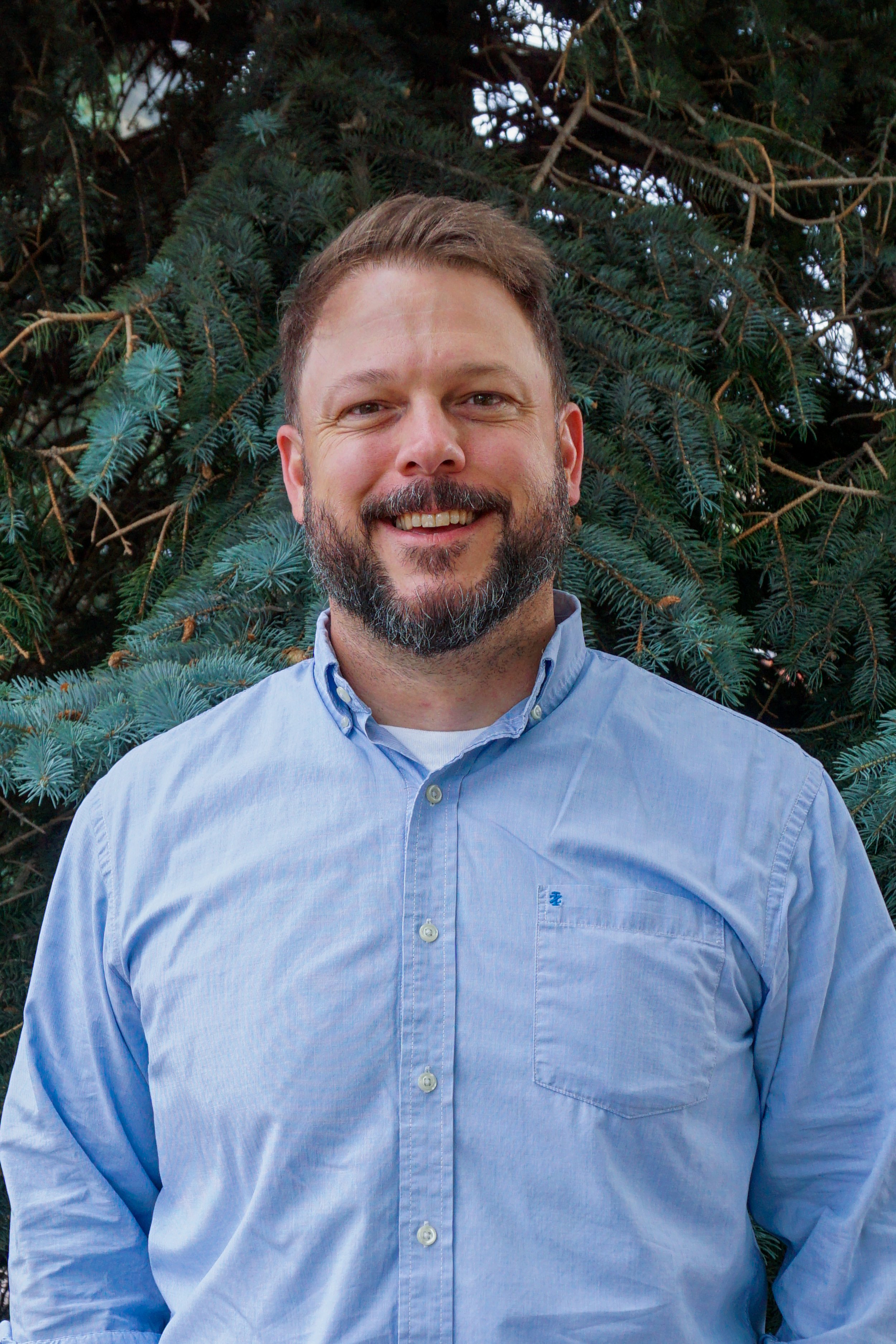 A smiling man with a beard wearing a light blue button-up shirt standing outdoors in front of a pine tree.