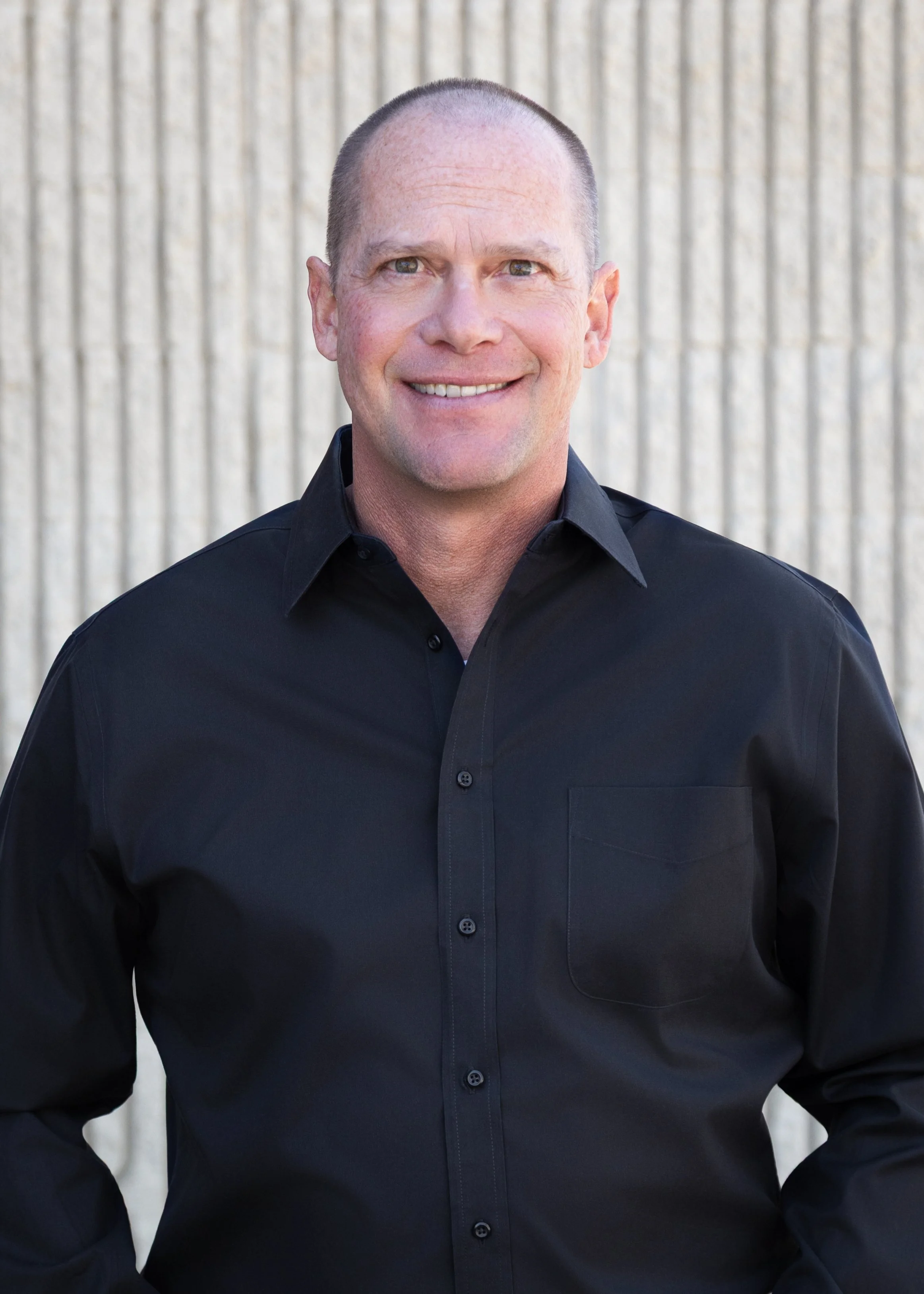 A middle-aged man with a clean-shaven face, short hair, and a smile, wearing a black button-up shirt, standing in front of a beige textured concrete wall.