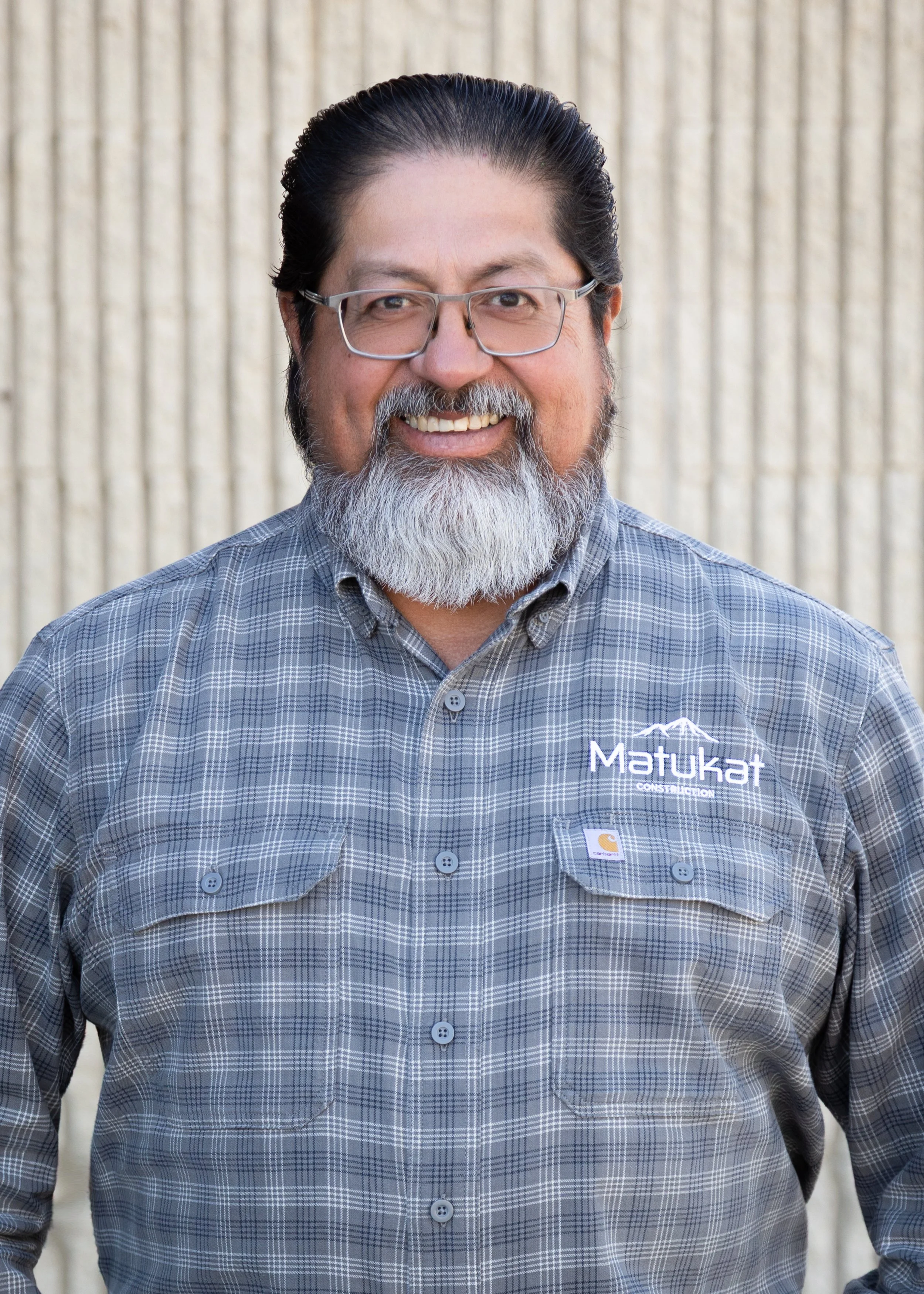 A middle-aged man with glasses and a beard, smiling, wearing a gray plaid button-up shirt with company logos, standing in front of a striped beige wall.