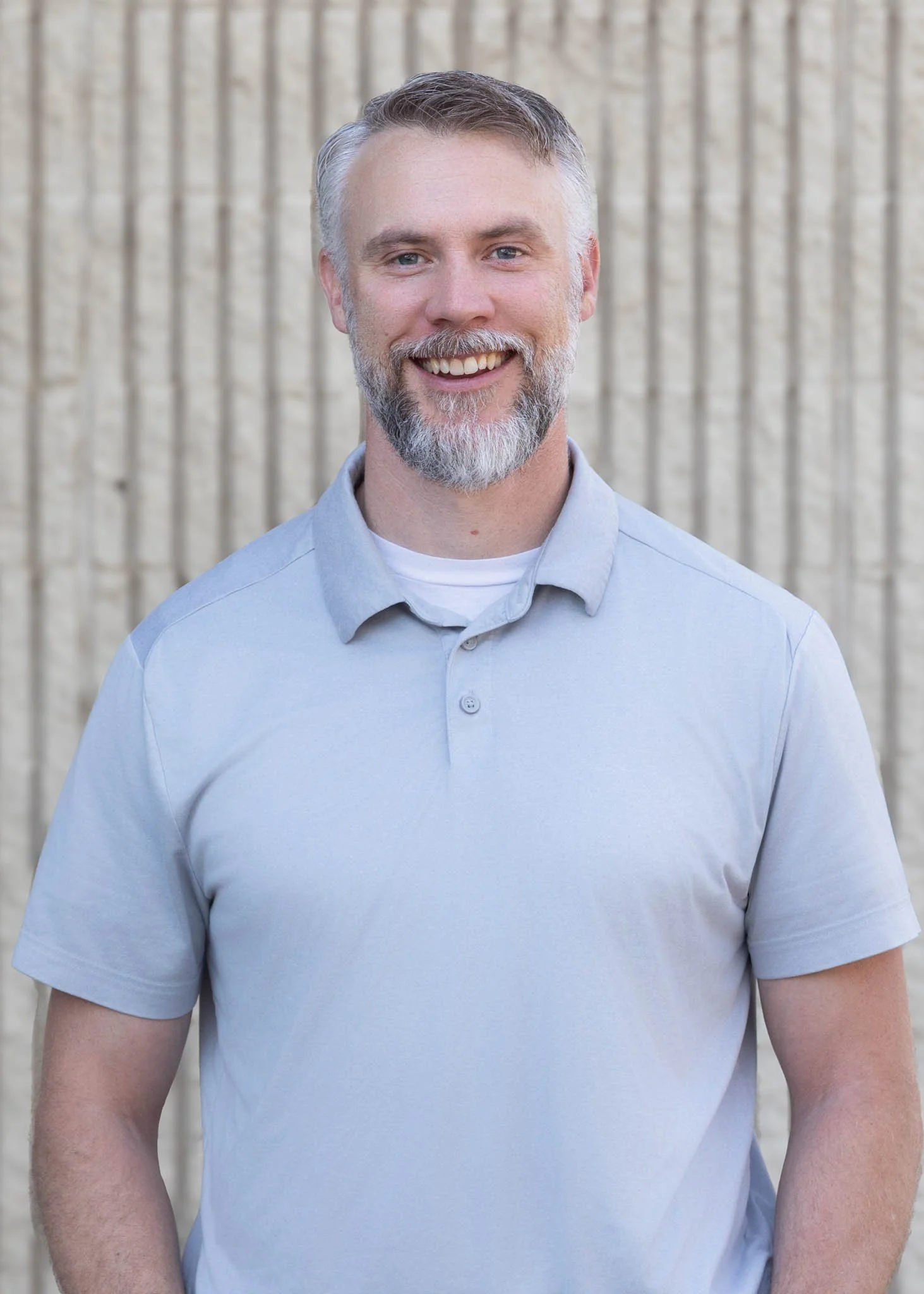 A smiling man with short gray hair and a beard, wearing a blue checkered shirt, standing outdoors in front of a large evergreen tree.