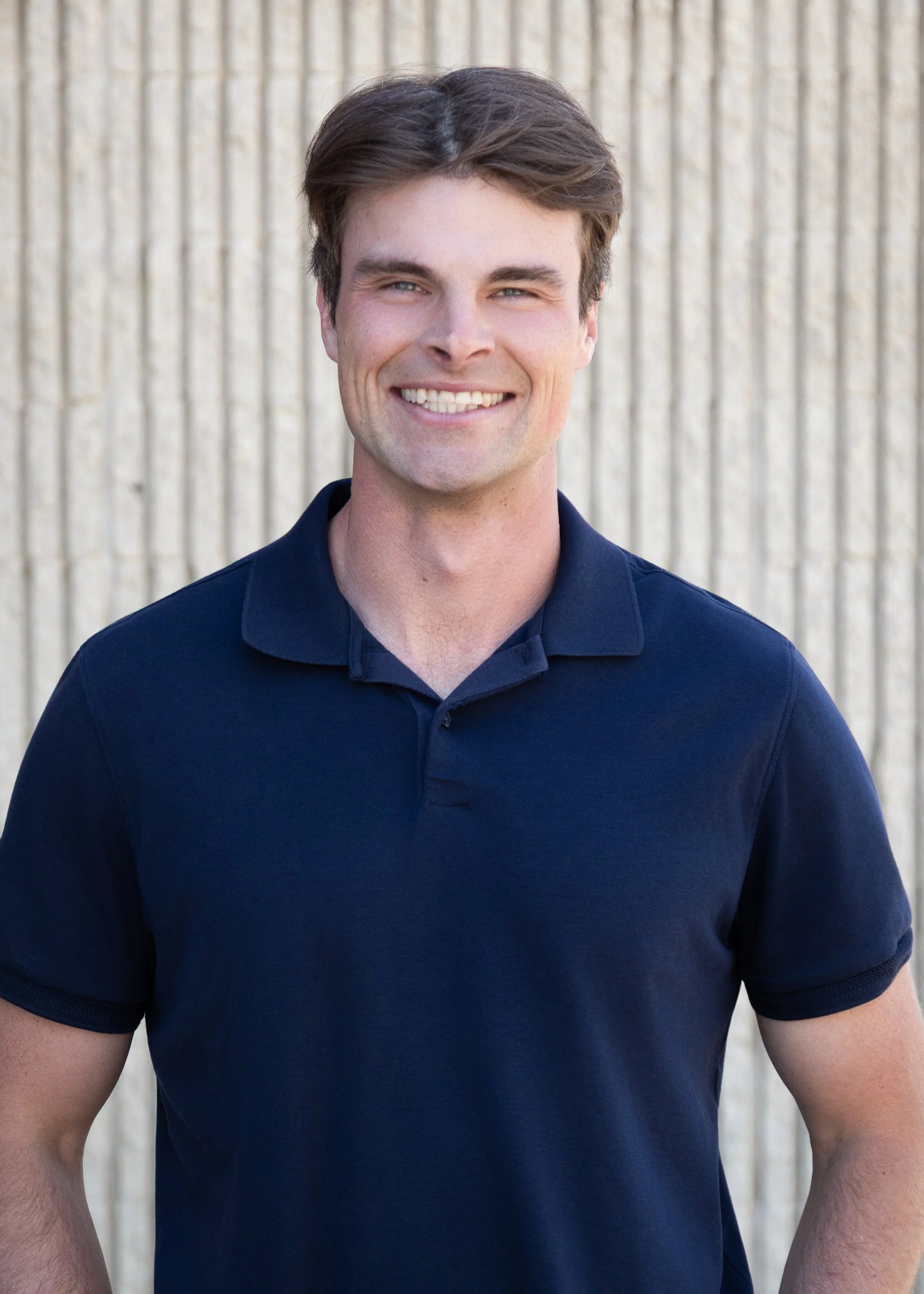 Head and upper body of a smiling man wearing a navy blue polo shirt, standing outdoors in front of a vertical beige panel wall.
