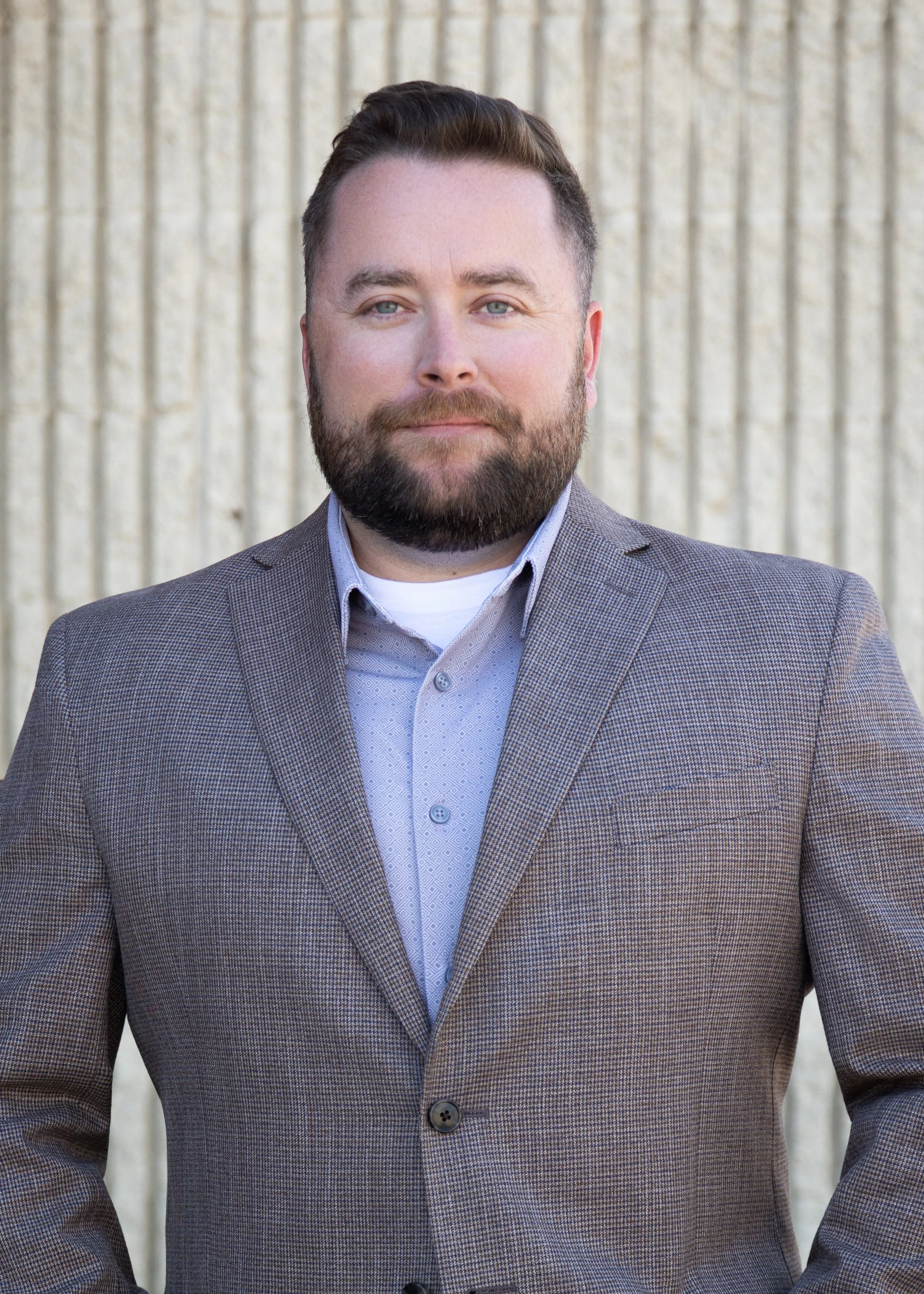 A man in a checkered brown blazer and a light patterned shirt standing in front of a beige corrugated wall.