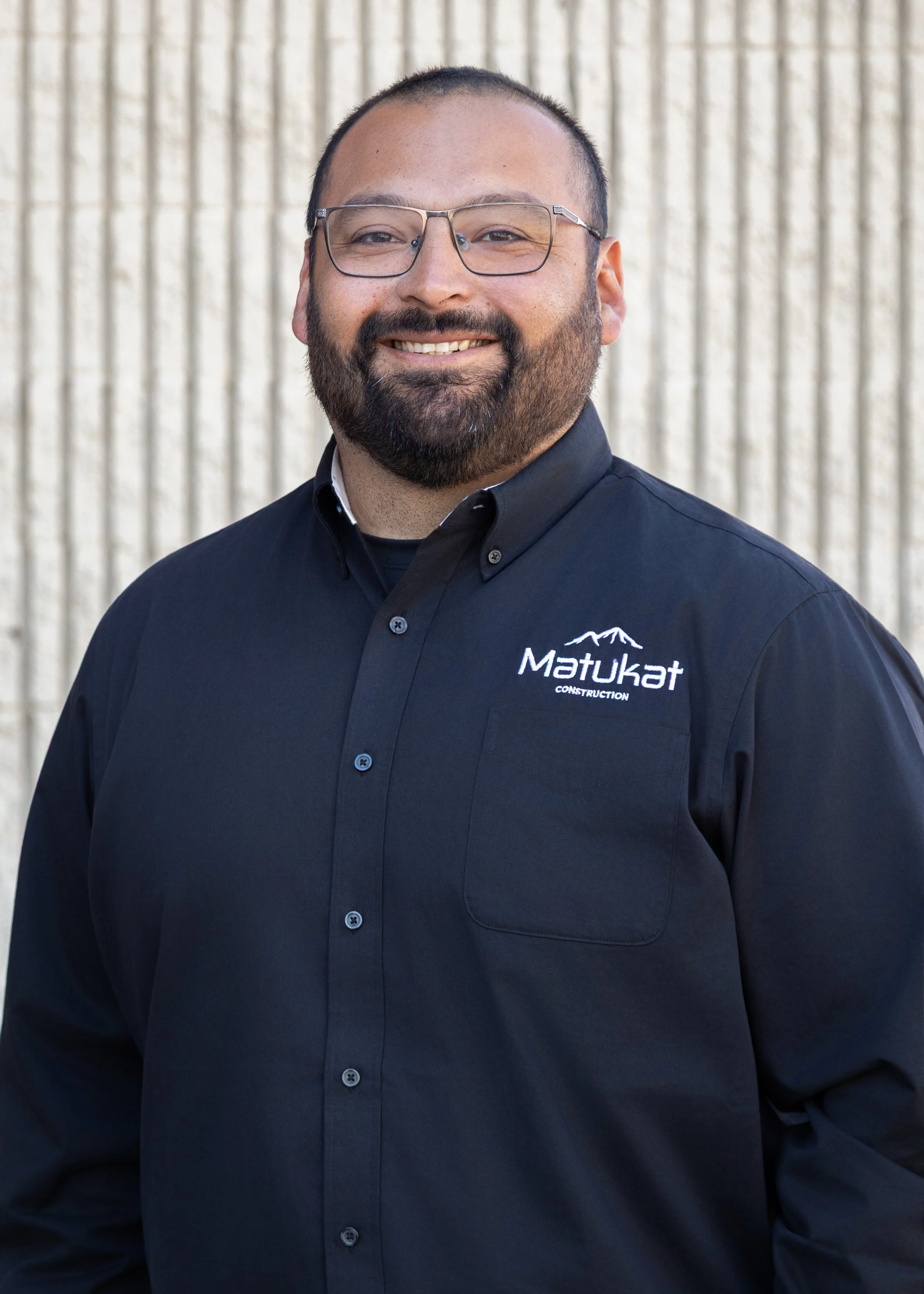 A man with glasses and a beard smiling, wearing a dark shirt with 'Matukat Construction' logo, standing against a beige textured wall.