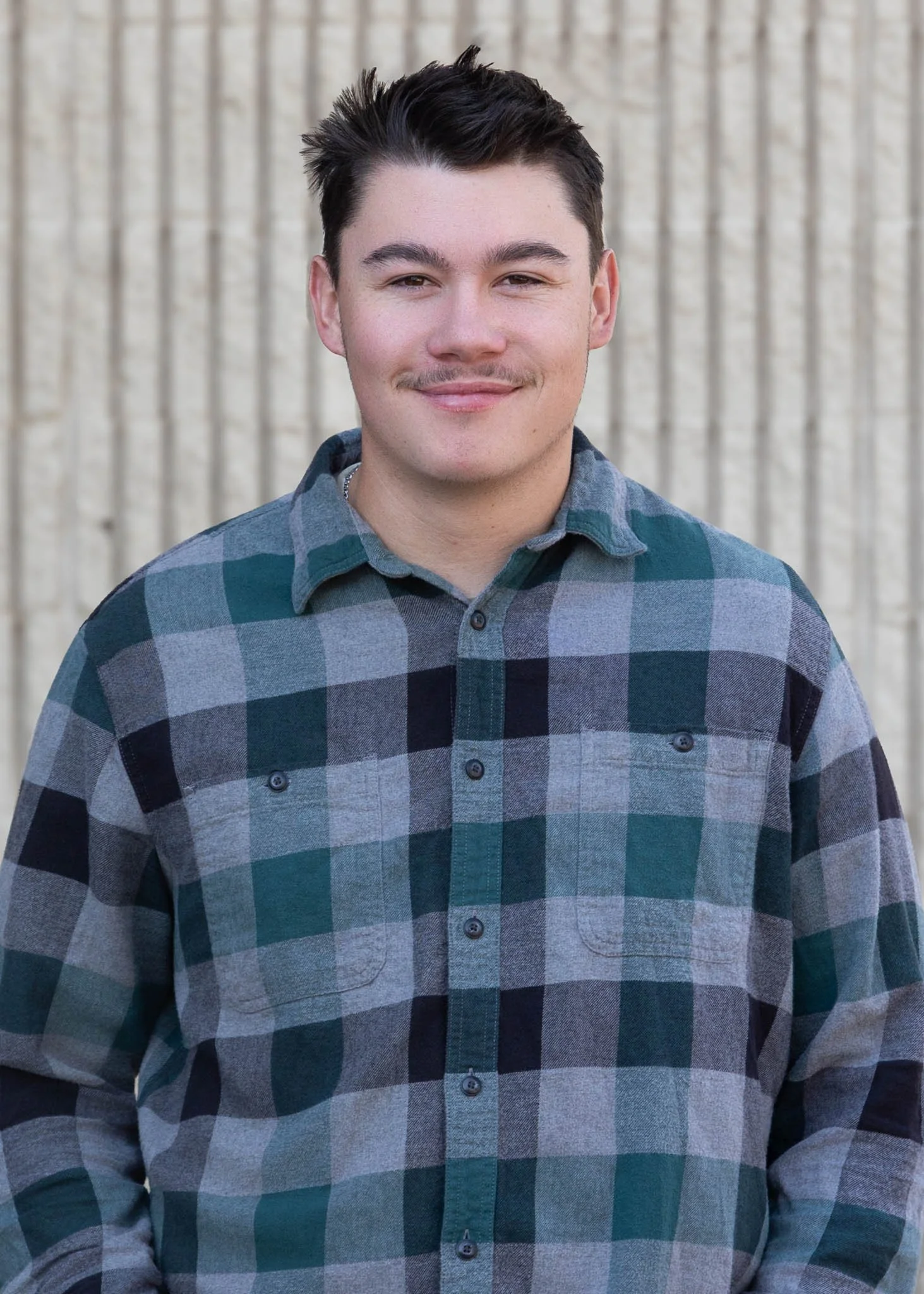 A young man standing outdoors in front of a green pine tree, wearing a plaid shirt, smiling at the camera.