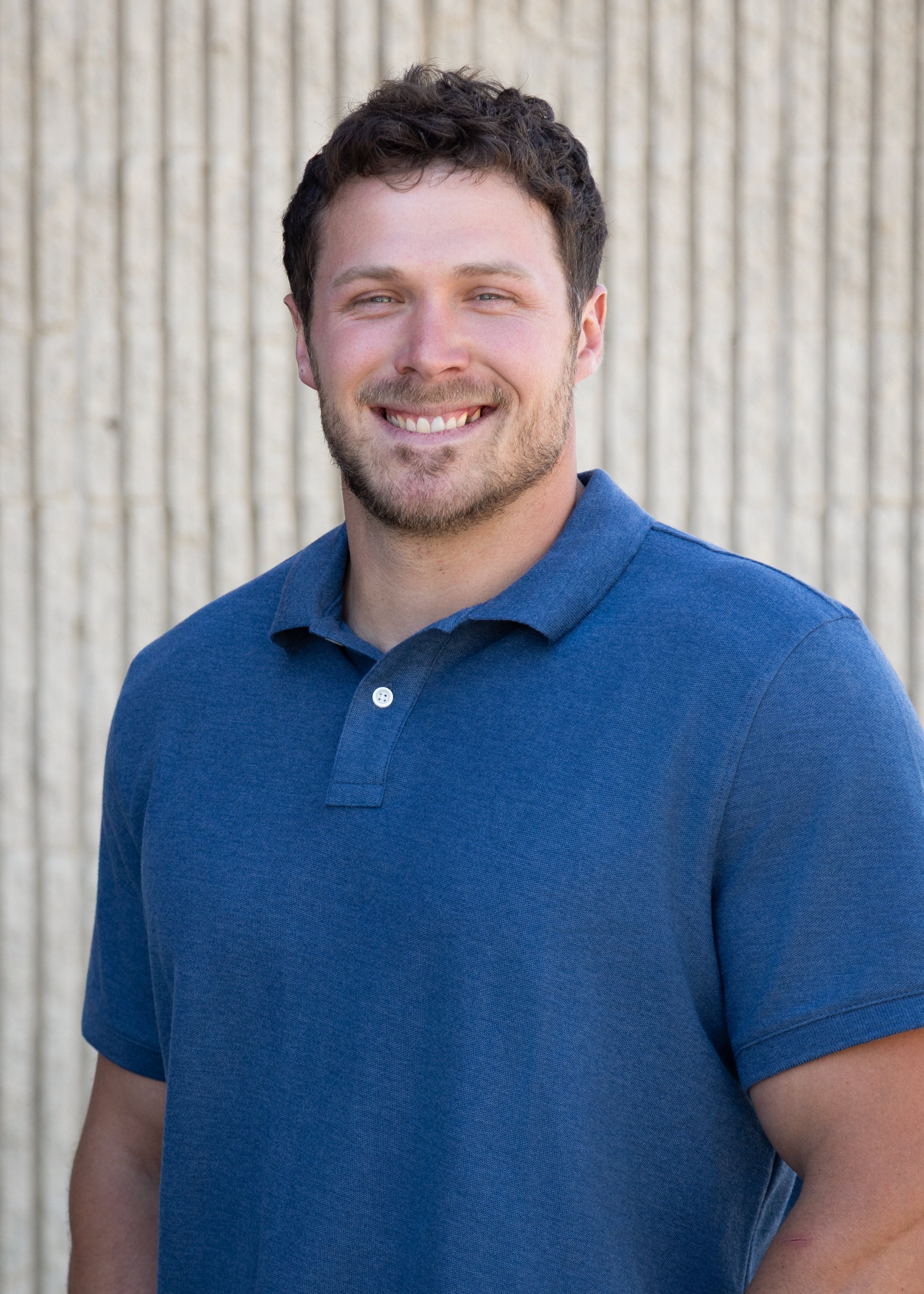 A young man with short, curly brown hair, light complexion, and blue eyes, smiling and wearing a blue polo shirt, standing outdoors in front of a textured, beige wall.