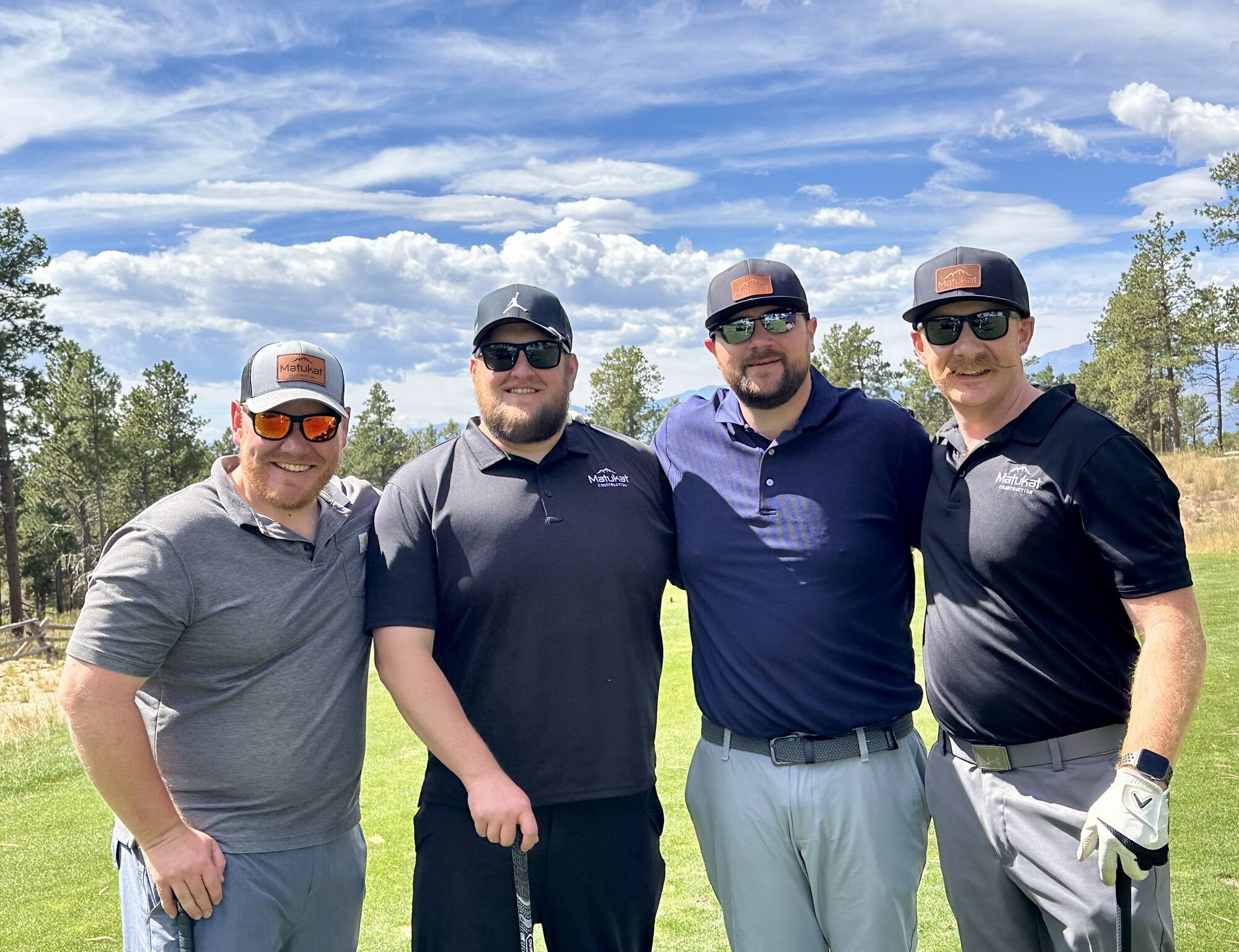 Four men standing outdoors on a golf course, smiling and dressed in golf attire, with trees and a partly cloudy sky in the background.