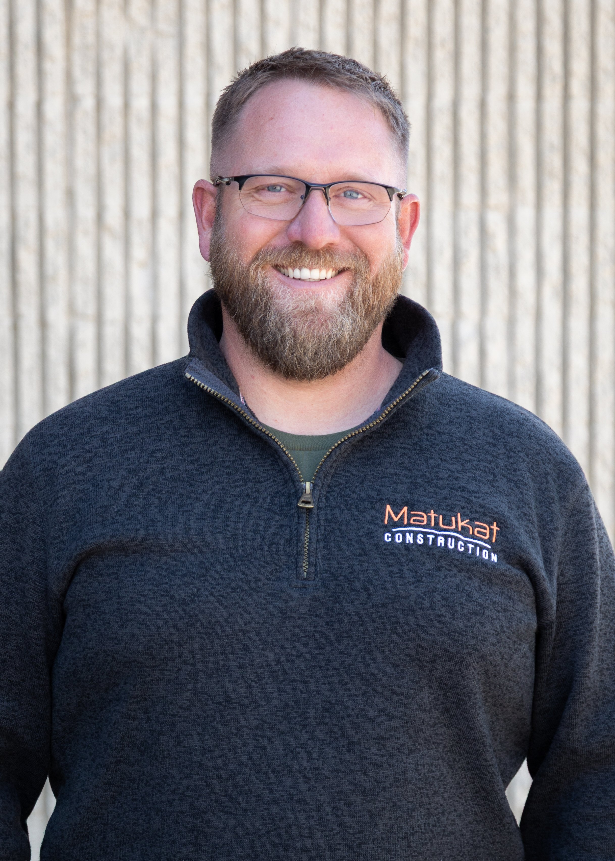 A smiling man with glasses and a beard wearing a dark gray zip-up jacket with a company logo that reads 'Matukat Construction' visible on the chest. Background is a textured beige wall.