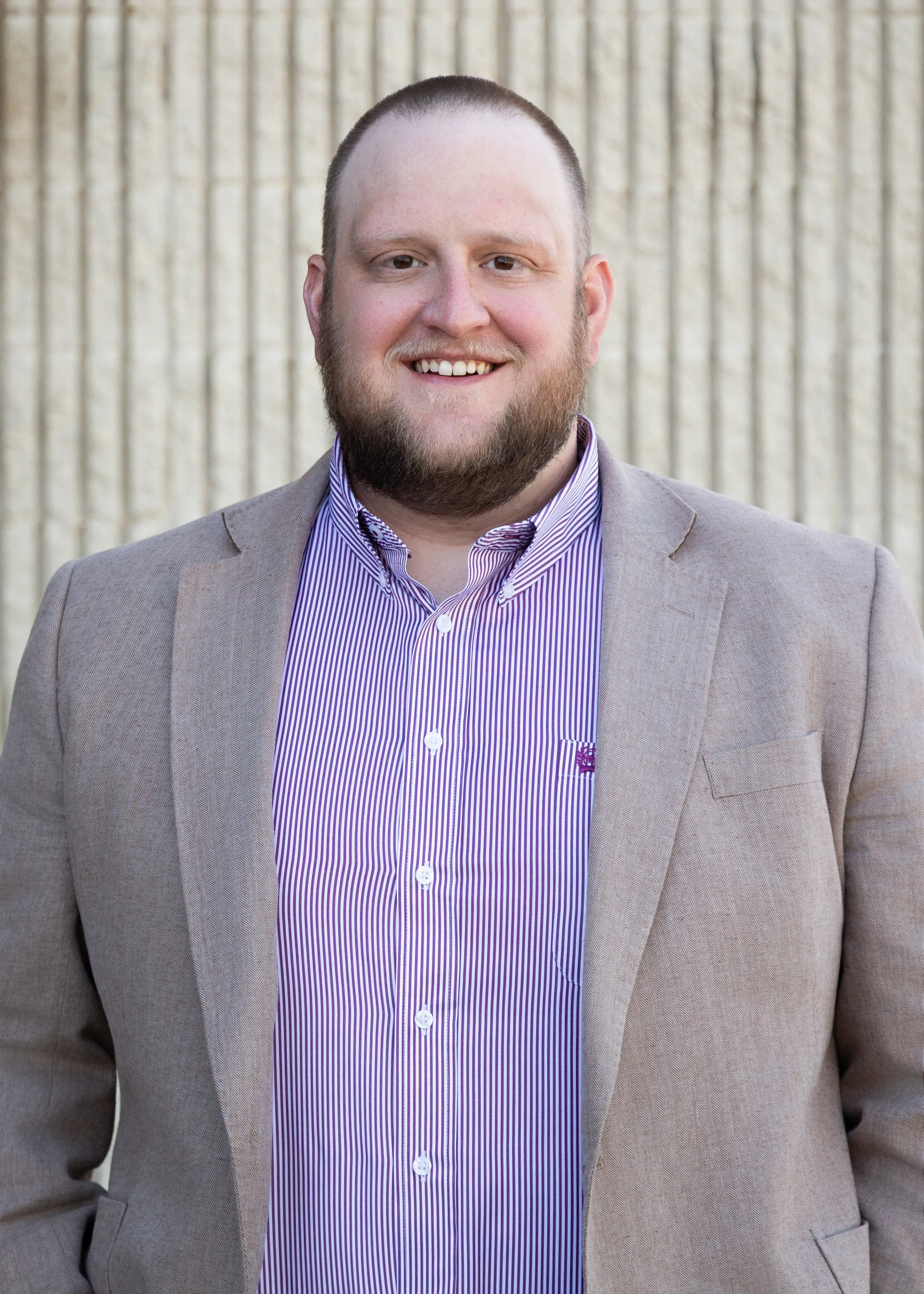 A smiling man with a beard and short hair wearing a gray blazer and a purple and white striped dress shirt, standing outdoors with a beige textured wall in the background.