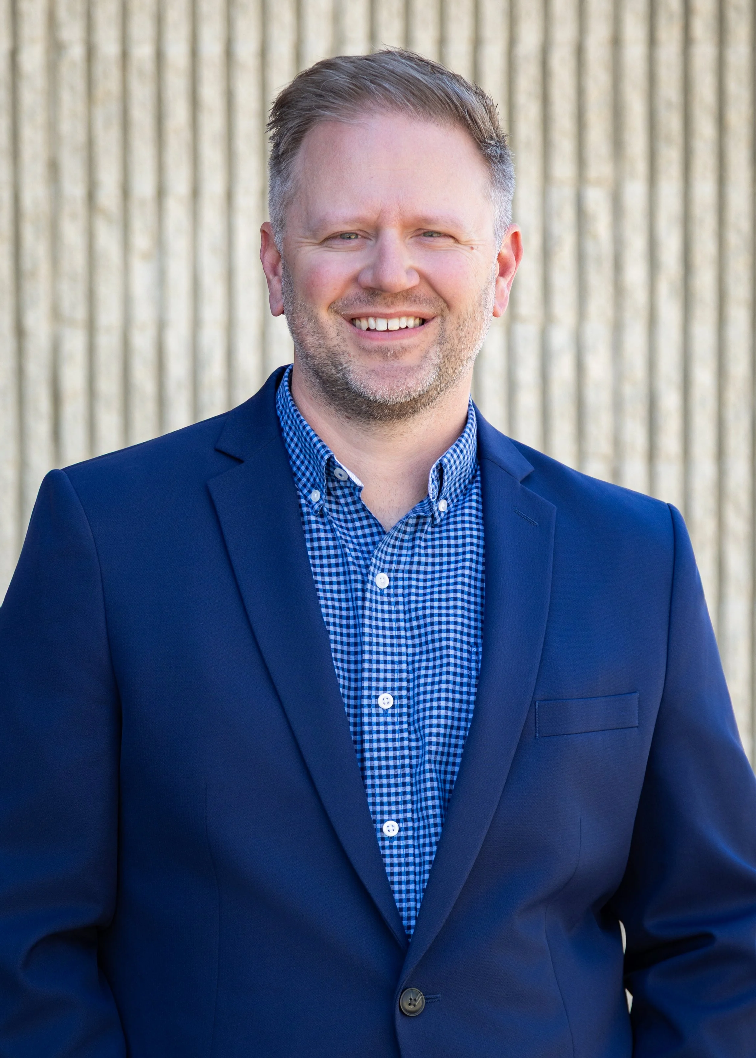 Headshot of a middle-aged man with short, slicked-back hair, wearing a dark blue blazer over a blue checkered shirt, smiling in front of a blurred outdoor background.