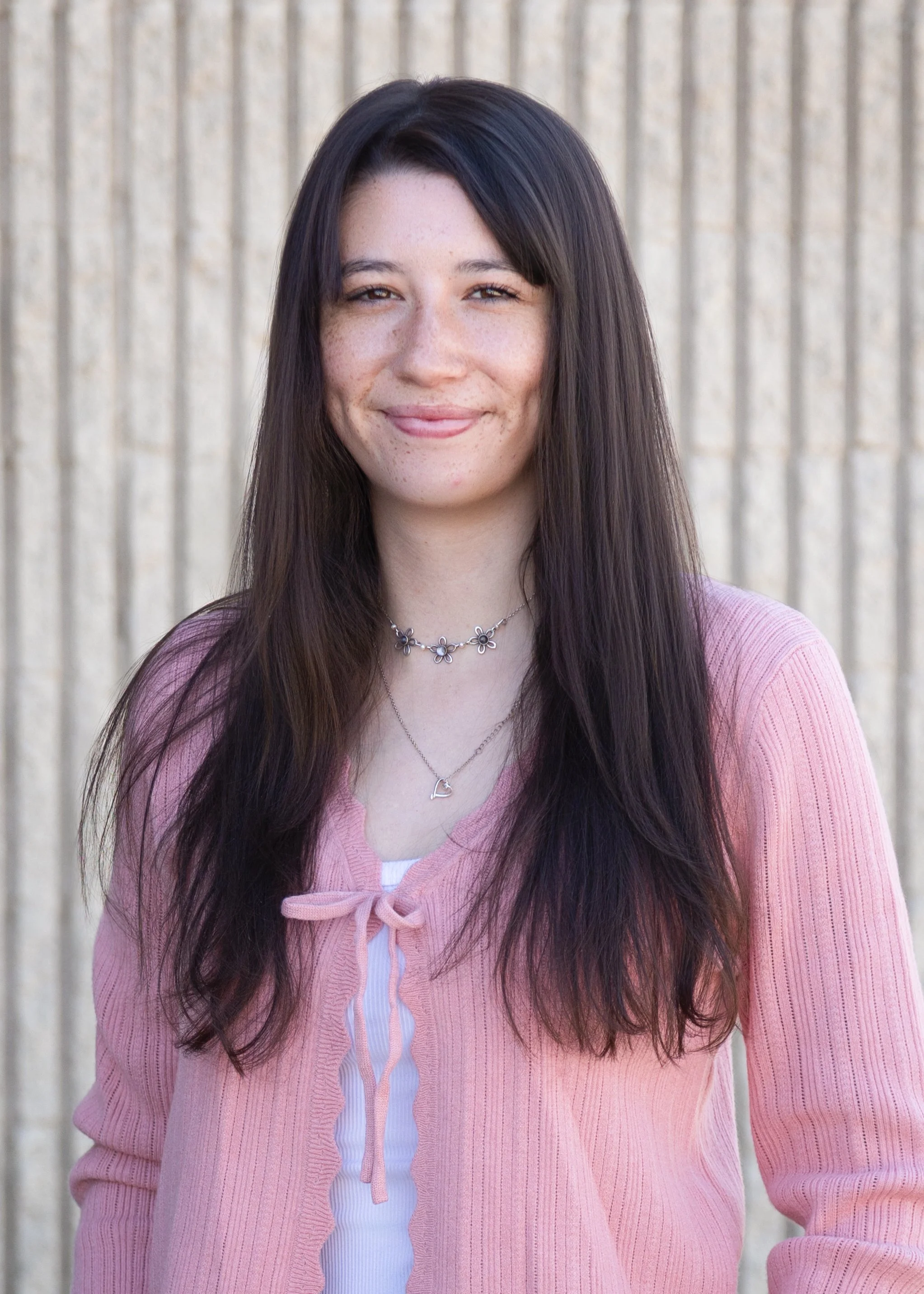 A young woman with long dark hair, smiling, standing outdoors in front of a beige vertical striped wall, wearing a pink cardigan over a white tank top, with layered necklaces.