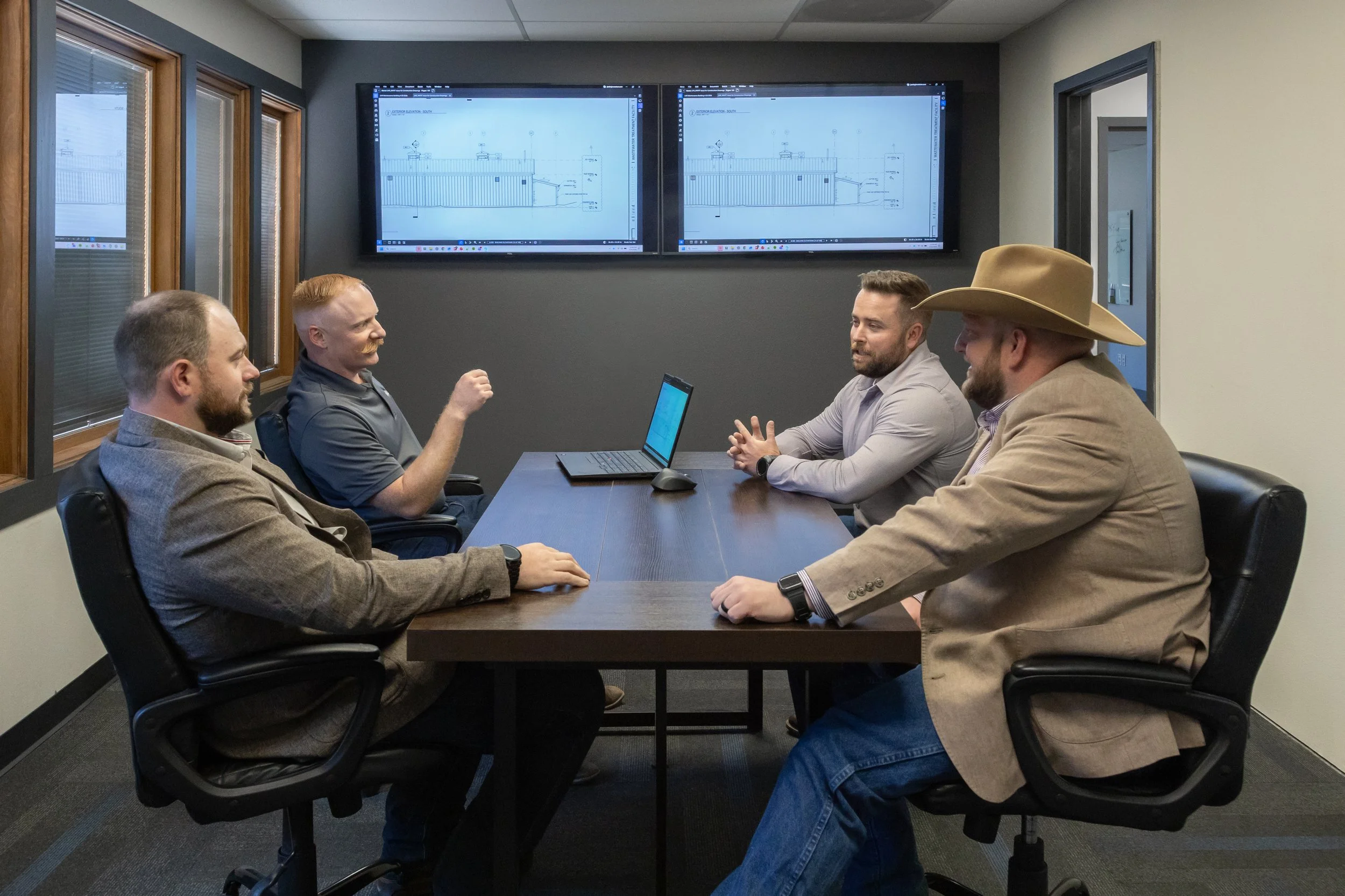 A group of four men sitting around a conference table in a meeting room, discussing blueprints displayed on two large screens behind them.