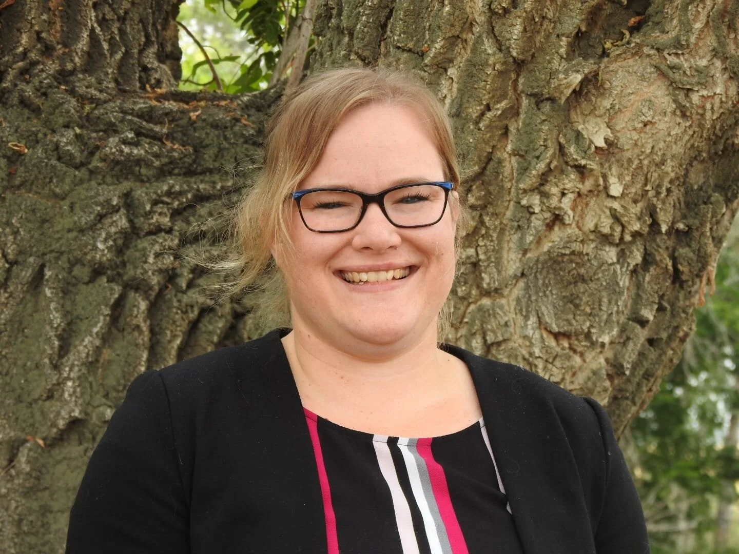 A woman with glasses and red hair smiling outdoors in front of a tree.