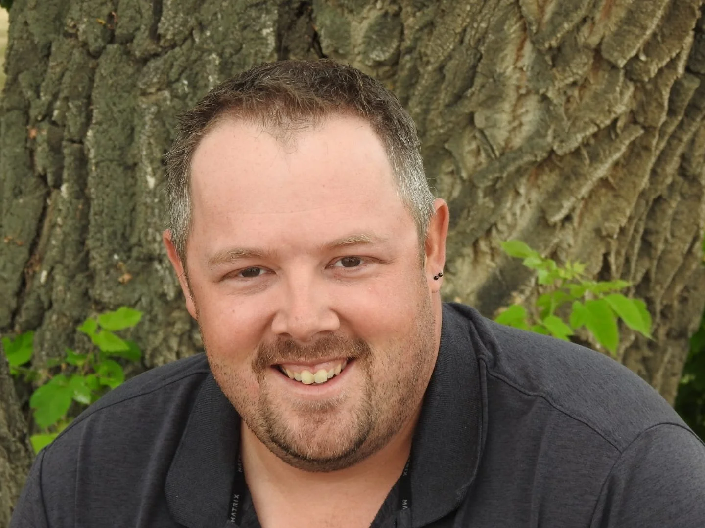 A smiling man with light skin, short brown hair, and a beard, sitting outdoors in front of a large tree trunk with green leaves.