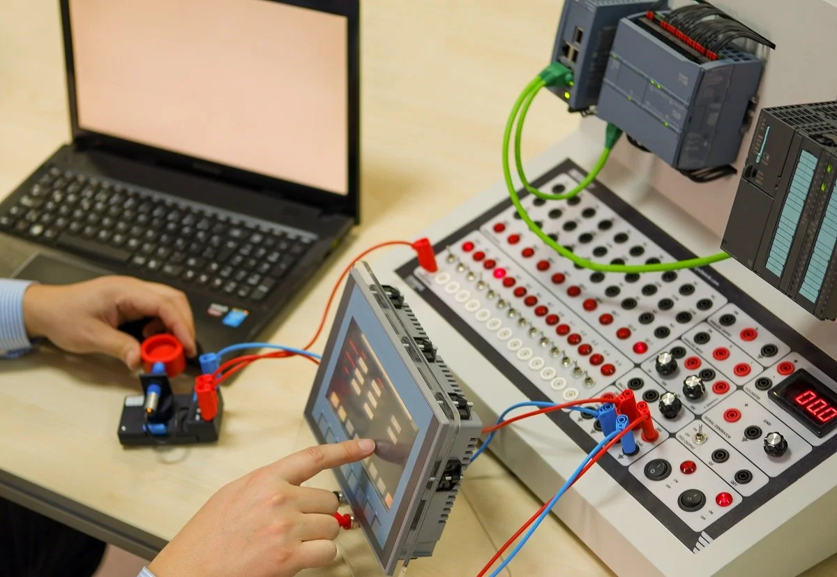 An engineering setup with a laptop, a control panel with red and black switches and knobs, a breadboard connected to a touchscreen controller, and programmable logic controllers, likely for automation or robotics testing.