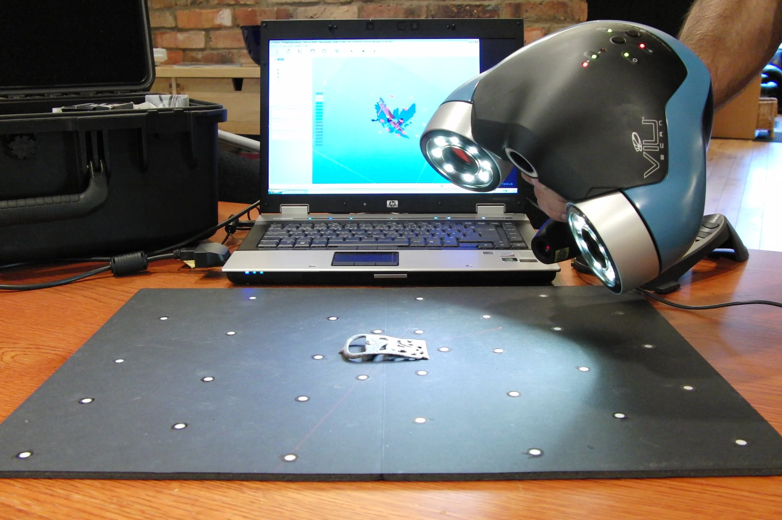A laptop displaying a colorful 3D digital model on its screen, with a black case partly open nearby, a modern helmet with LED lights and a logo resting on the laptop, and a small object on a black padded work surface on a wooden table, with a brick wall in the background.