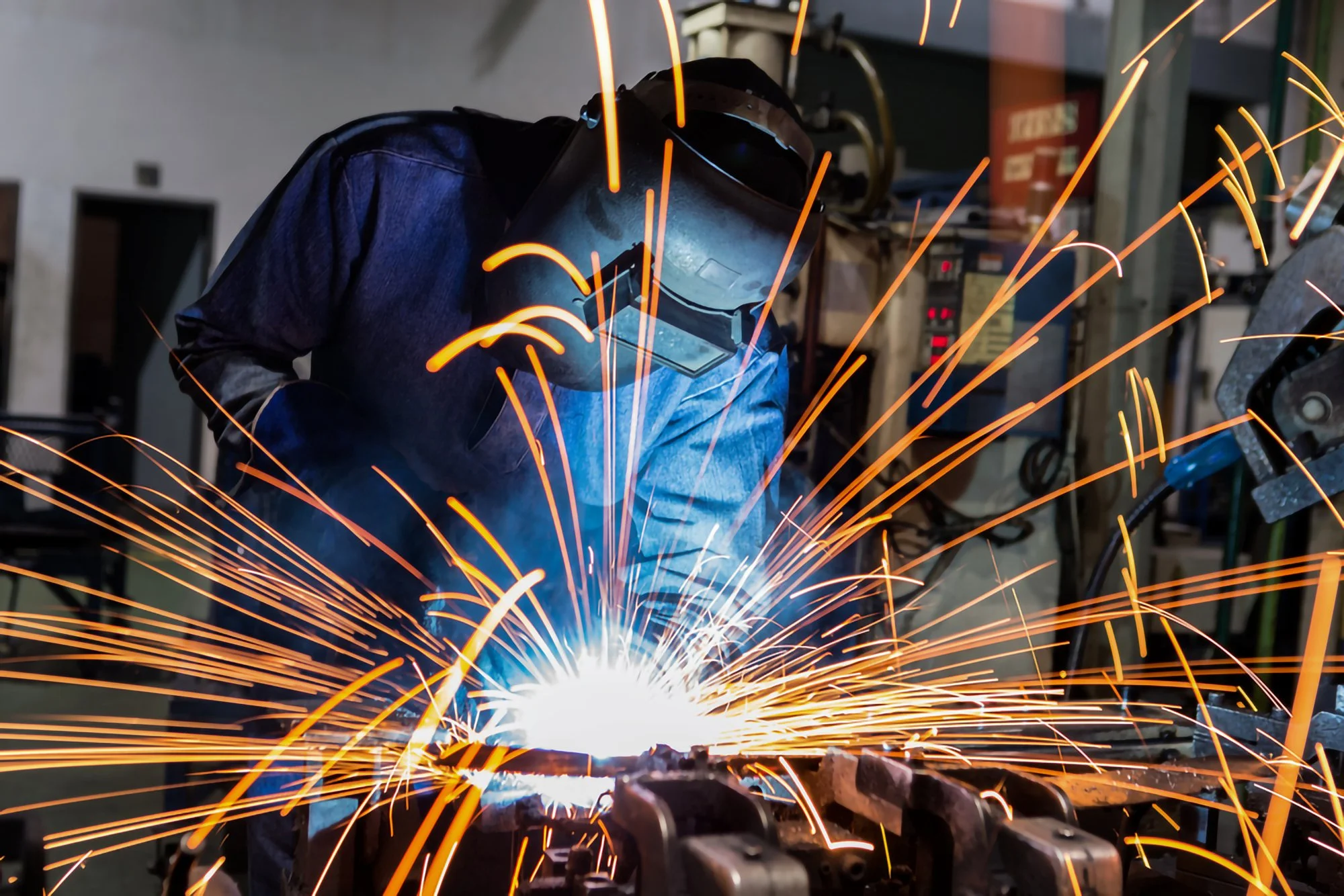 A person wearing protective welding gear, including a helmet and gloves, welding metal with sparks flying in a workshop setting.