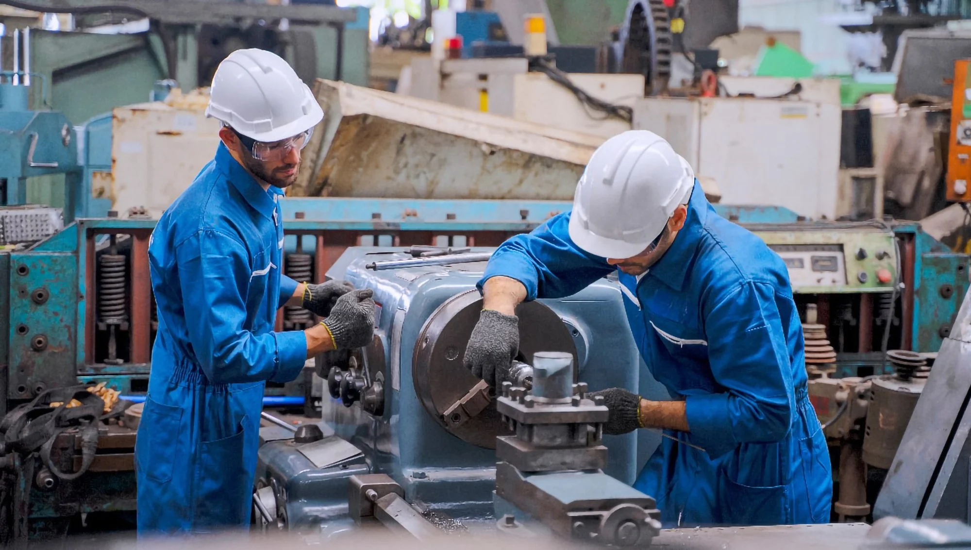 Two Breakthrough Industrial Solutions employees, wearing blue uniforms, white safety helmets, safety glasses, and gloves, perform preventive maintenance on a large industrial machine.