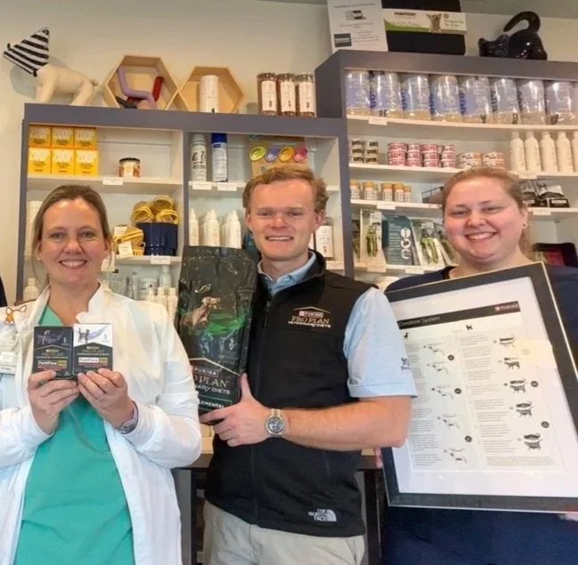 Three people in professional attire holding various pet health and nutrition products in a store or pharmacy setting, with shelves of pet supplies in the background.