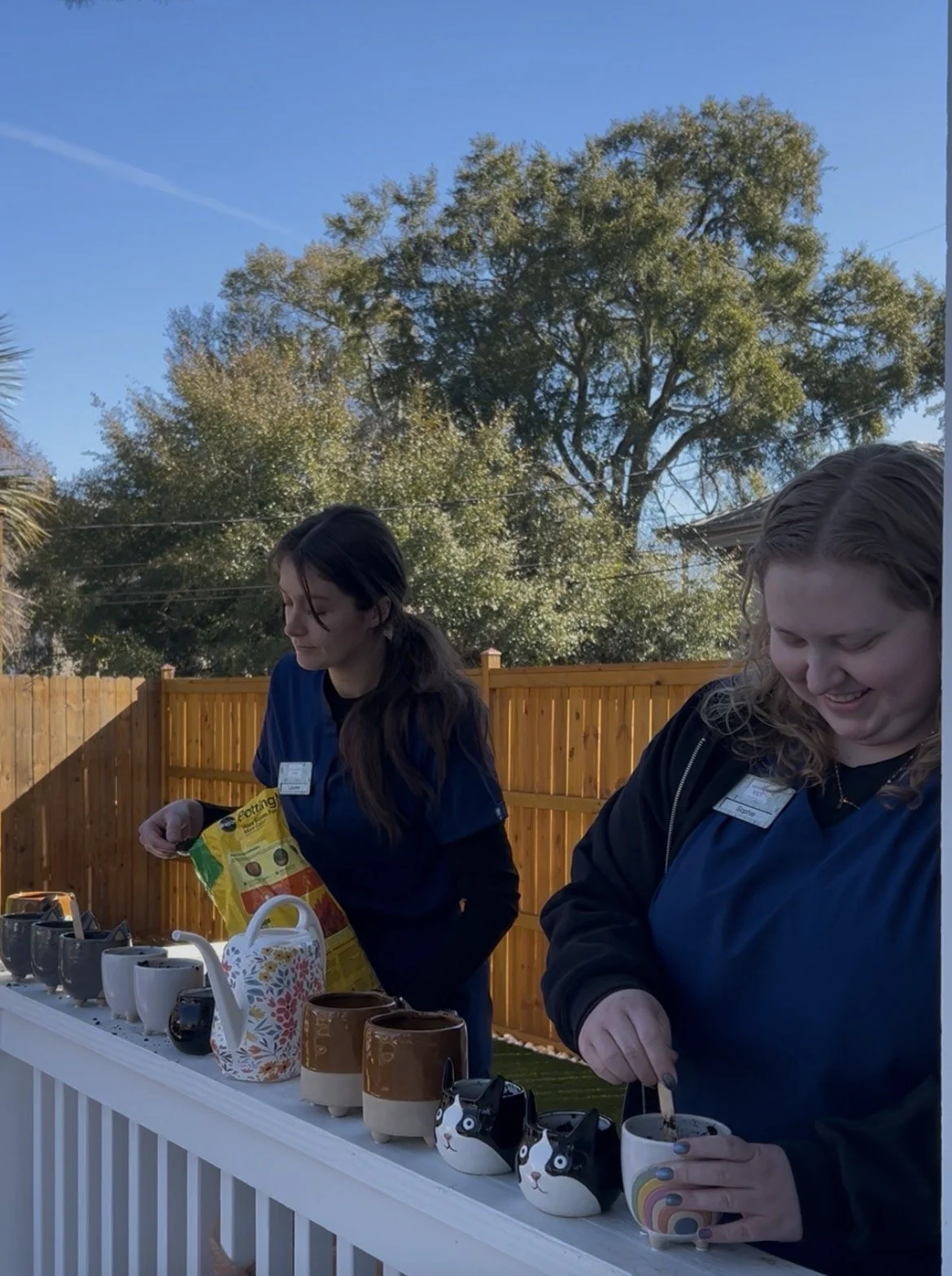 Two women in navy jackets standing at a table with various coffee mugs and a teapot outdoors in a backyard with a wooden fence, trees, and a clear blue sky.