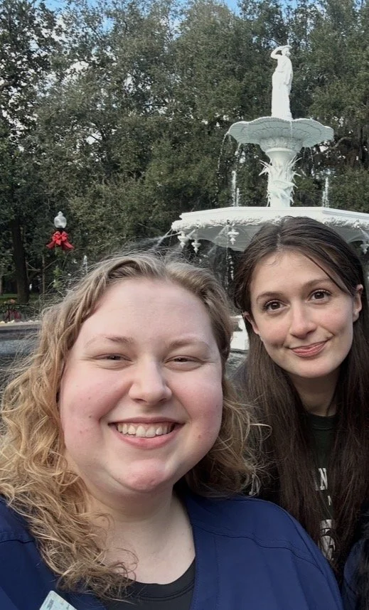 Two women smiling in front of a white fountain in Forsyth Park with a statue at the top, outdoors with trees in the background.
