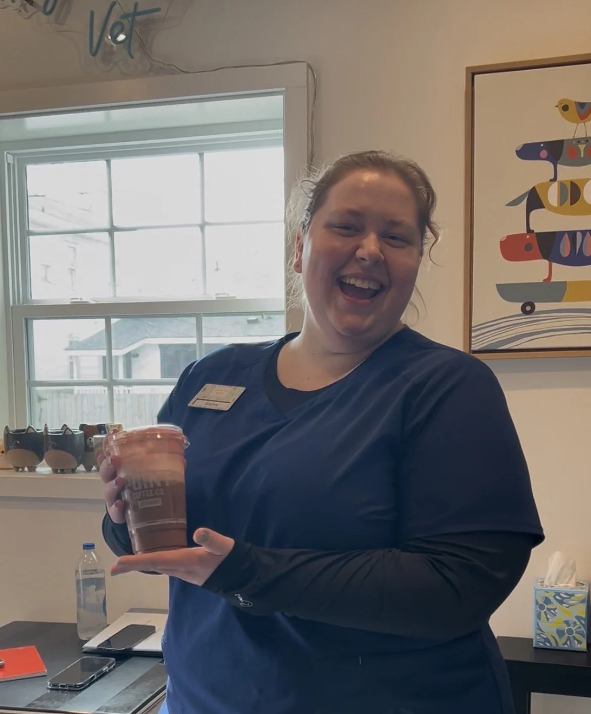 A woman in scrubs holding a large iced coffee in a room with artwork and a window.