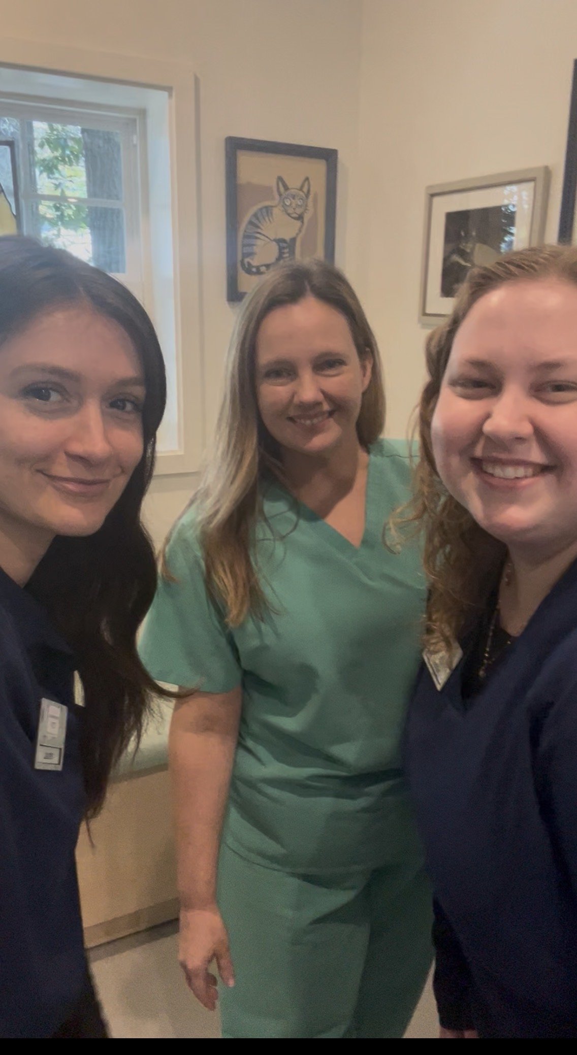 Three women smiling and posing for a selfie in an indoor setting, with framed pictures, including a cat, and a window in the background.