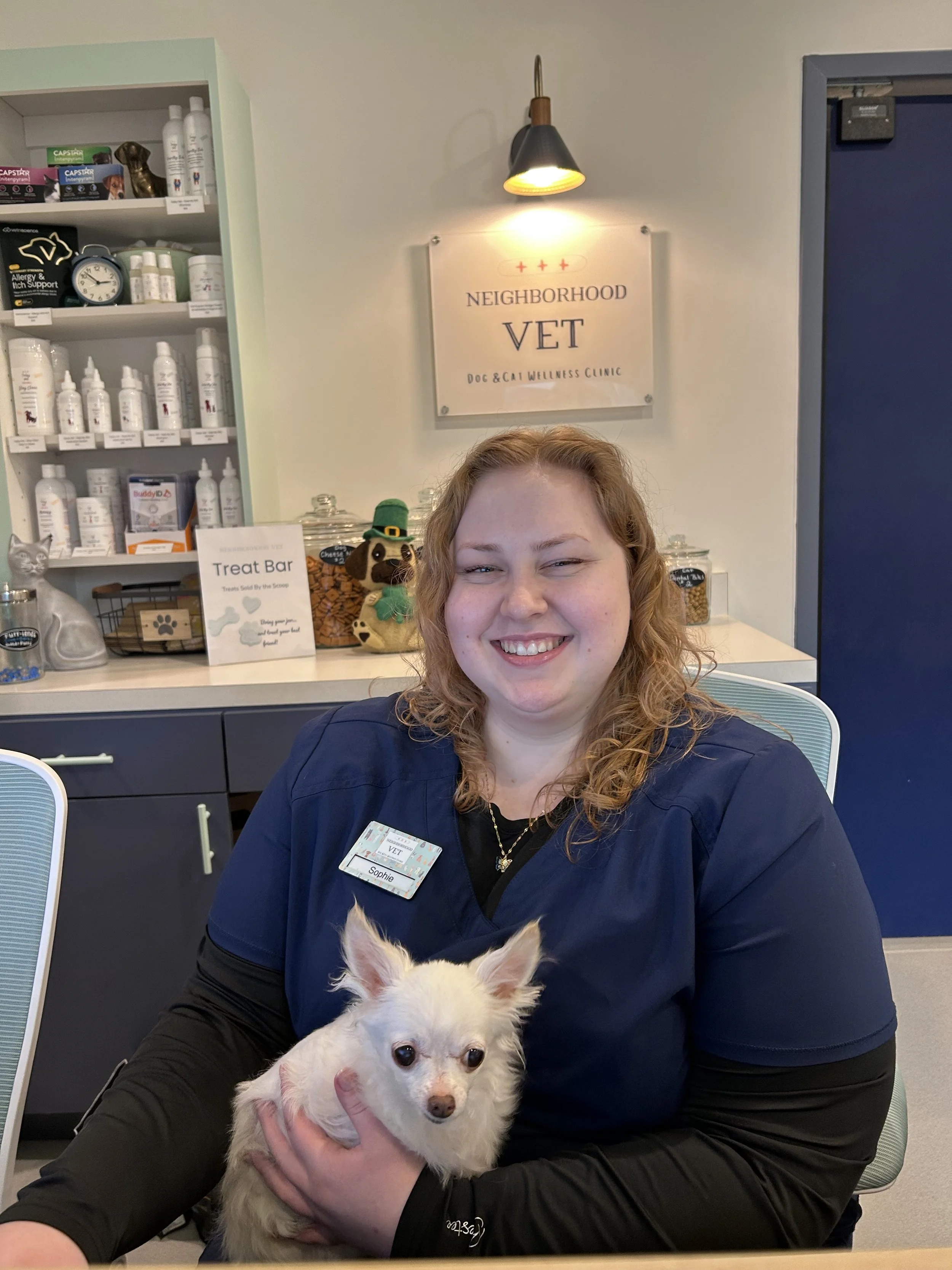 A woman in navy scrubs smiling and holding a small white dog in a veterinary clinic. Behind her, there's a sign that reads 'Neighborhood VET' and shelves with bottles and pet supplies.