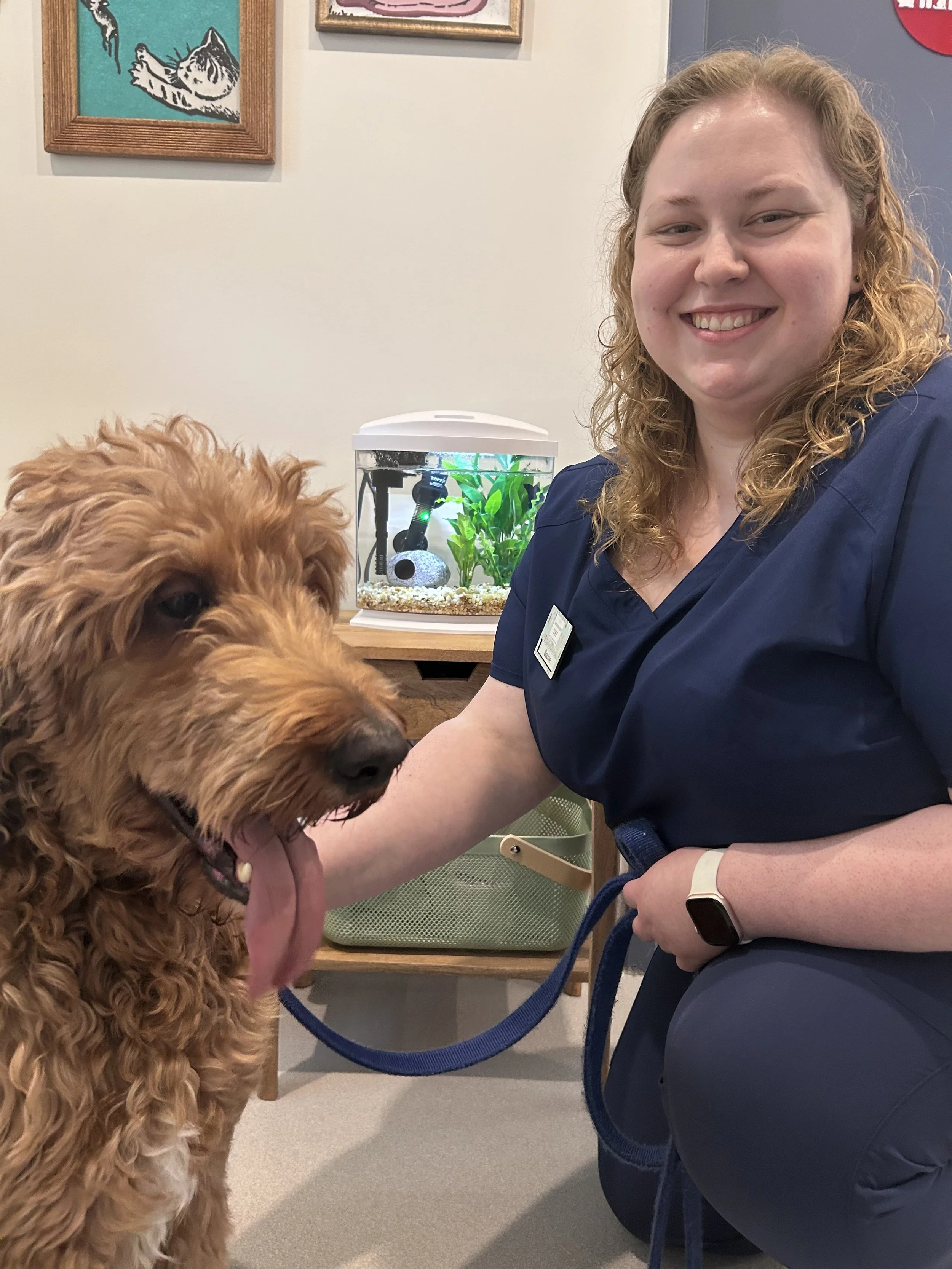A woman in navy scrubs smiling beside a curly-haired goldendoodle dog inside a veterinary office or pet care facility with a small aquarium in the background.