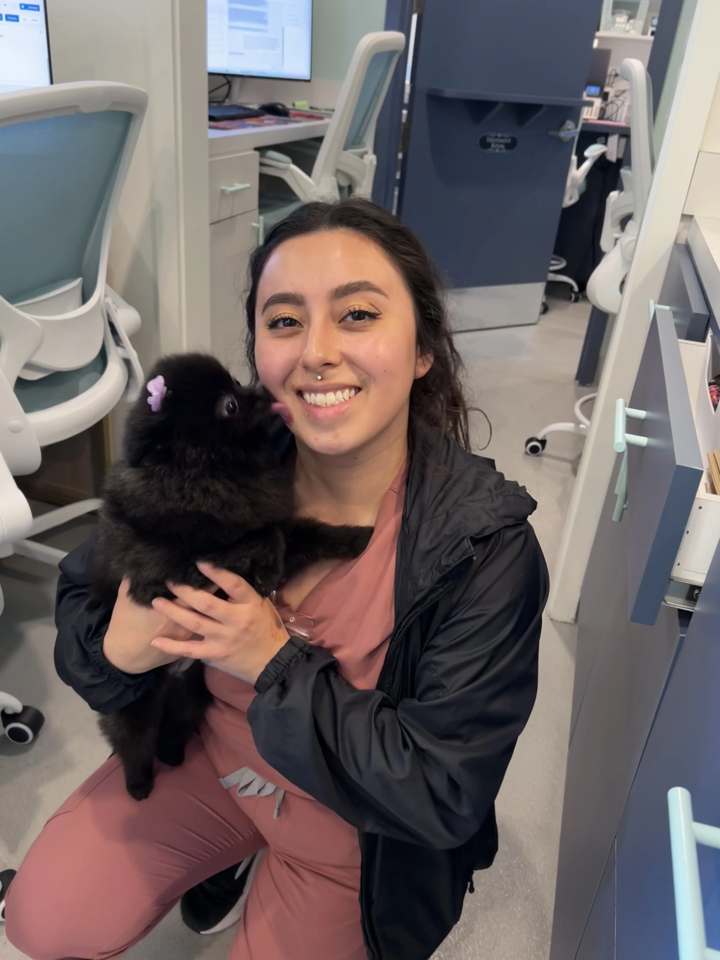 A young woman with dark hair, wearing a black jacket and pink pants, holding a black puppy with a pink bow on its ear, inside an office or medical setting with desks, chairs, and computer monitors in the background.