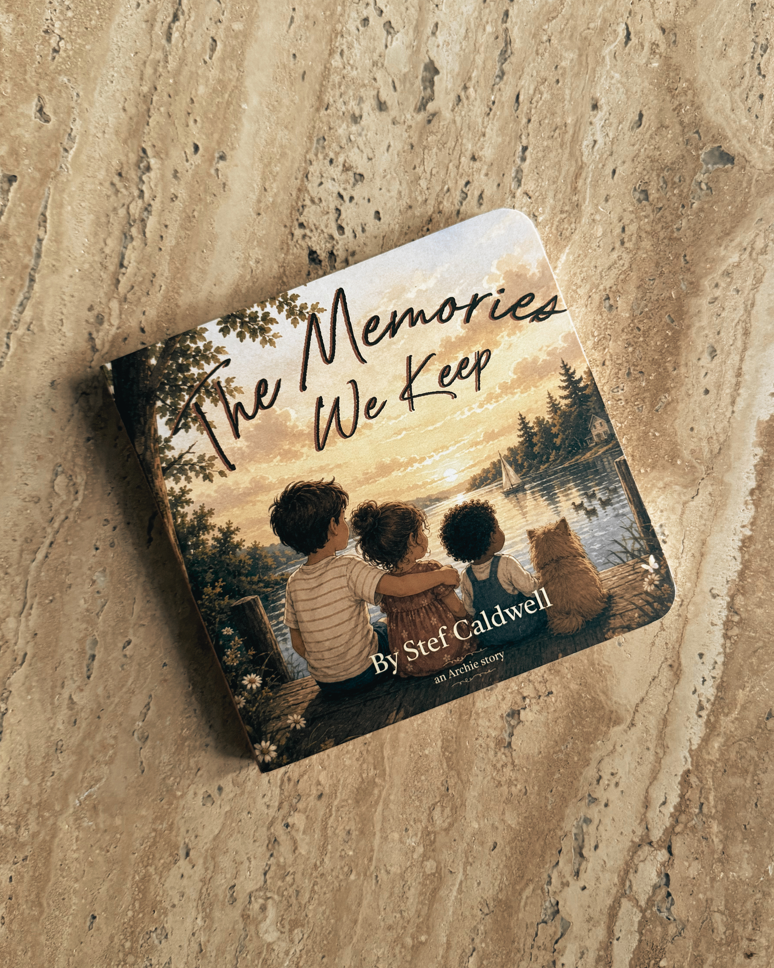 A book titled "The Memories We Keep" by Stef Caldwell resting on a textured beige surface. The cover illustration depicts three children sitting by a body of water, watching sailboats during a sunset.
